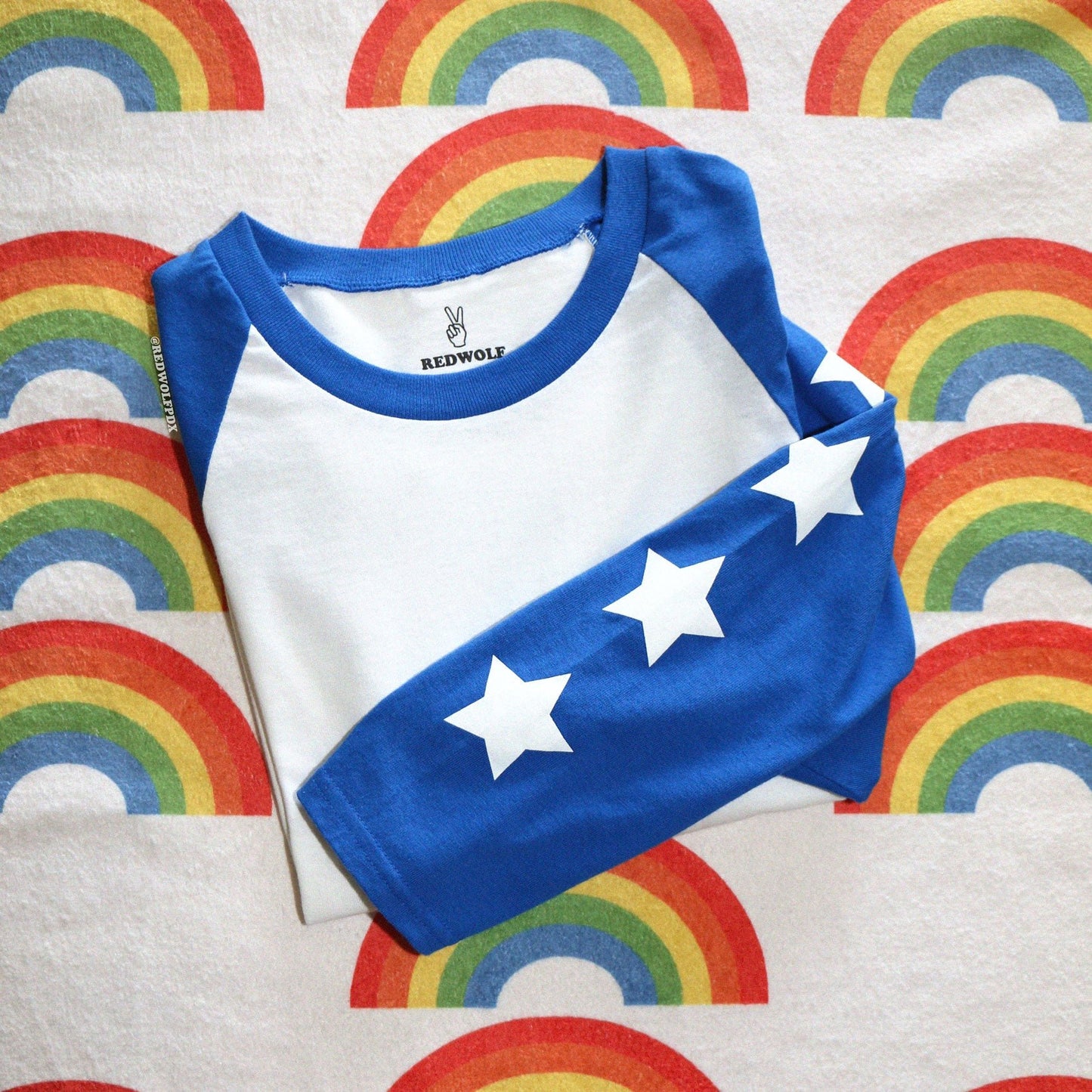 folded shirt with white stars screen printed down the bright blue sleeves of the raglan on a rainbow printed background