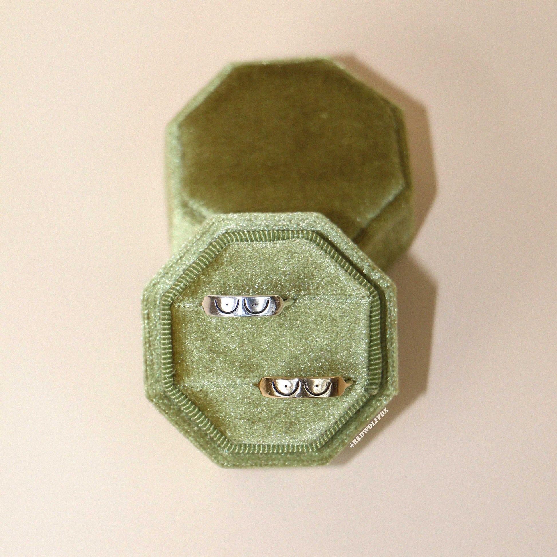 a sterling silver and a brass ring both with boob illustration etched across them displayed on a velvet green ring box against a beige background