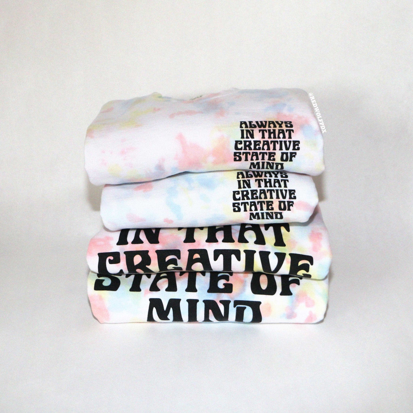 Stack of tie dye crewnecks with text 'ALWAYS ON THAT CREATIVE STATE OF MIND' screen printed on front and back on a white background