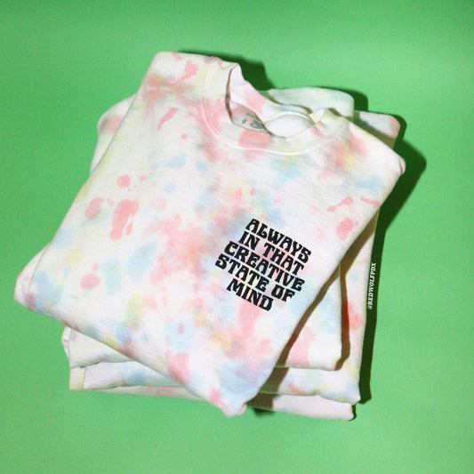 Stack of pastel tie-dye crewneck sweatshirts with text 'ALWAYS IN THAT CREATIVE STATE OF MIND' on a green background