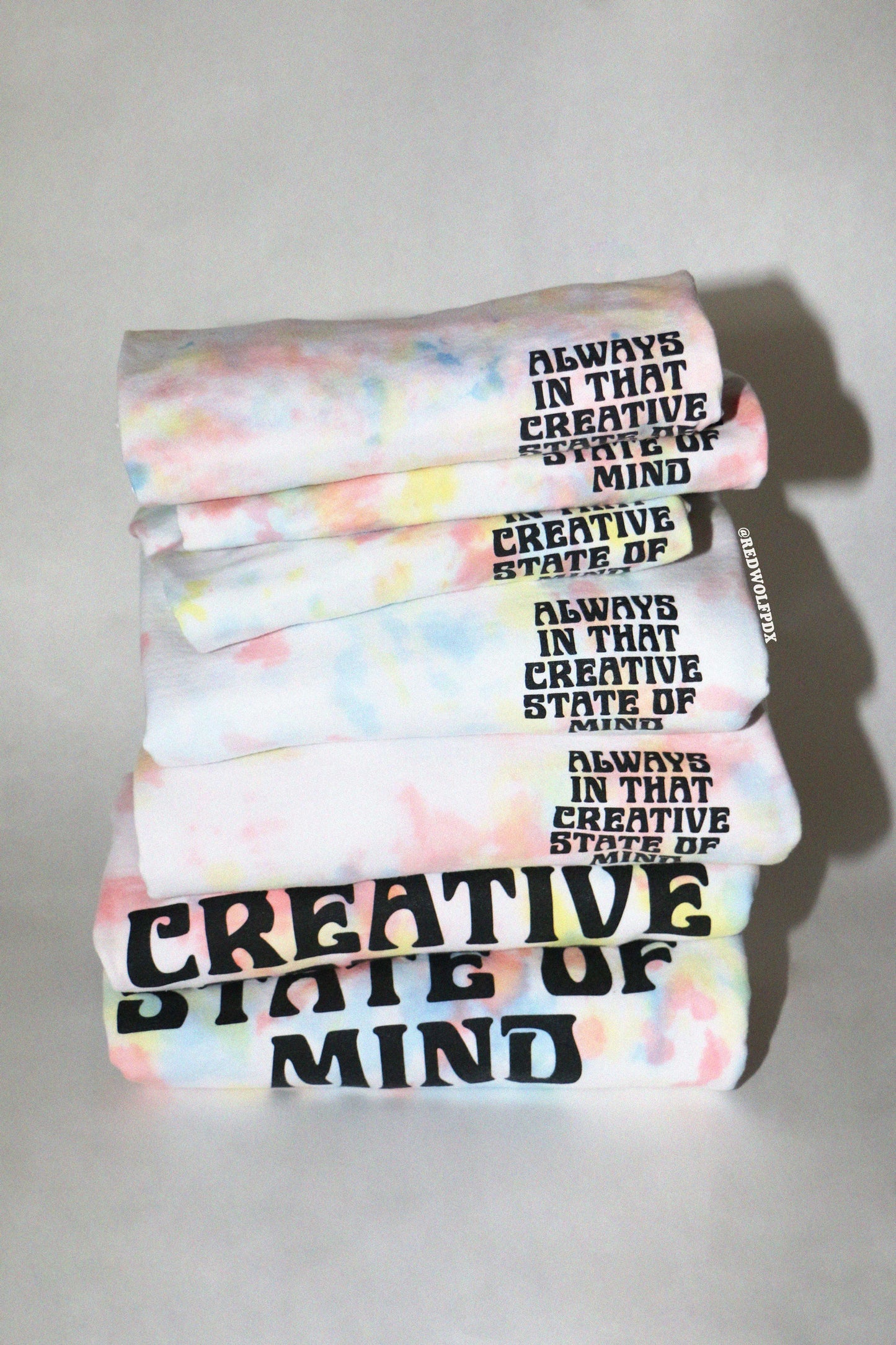 Stack of tie dye tees and crewnecks with the text 'Always in that creative state of mind' text on a white background