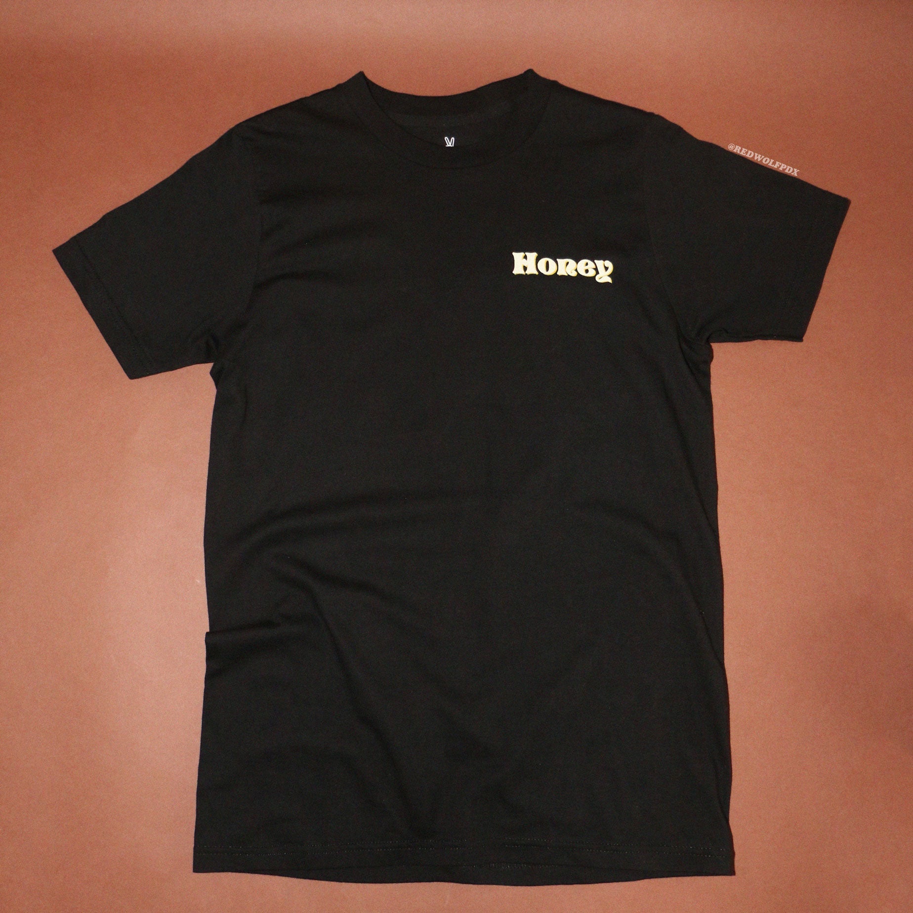 Black graphic tee with text 'Honey' in white ink shadowed with layer of yellow ink screen printed on corner of chest against a brown background