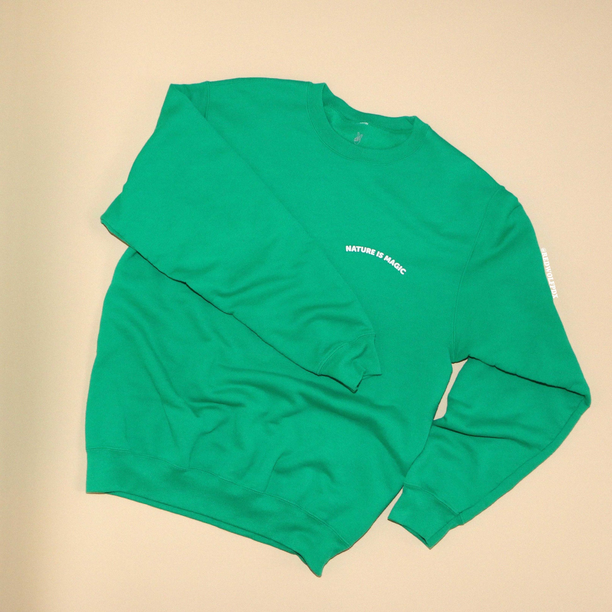 Green crewneck sweatshirt with text 'NATURE IS MAGIC' on front against beige backdrop