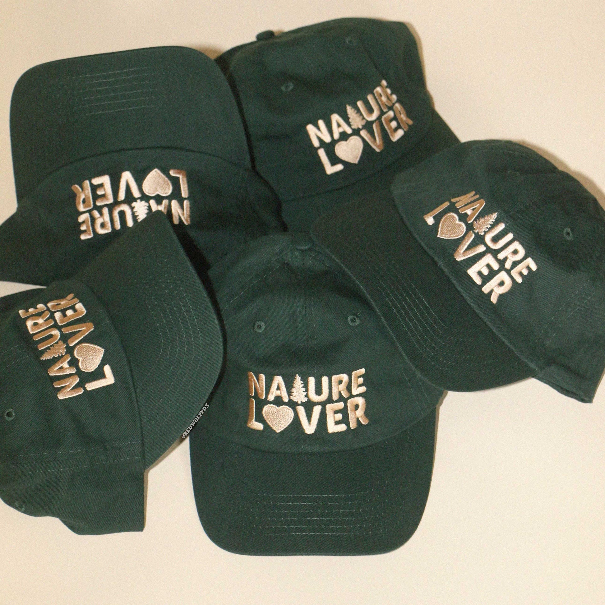 a pile of forest green baseball caps with the text 'NATURE LOVER' embroidered with beige thread across the front of them against a beige background