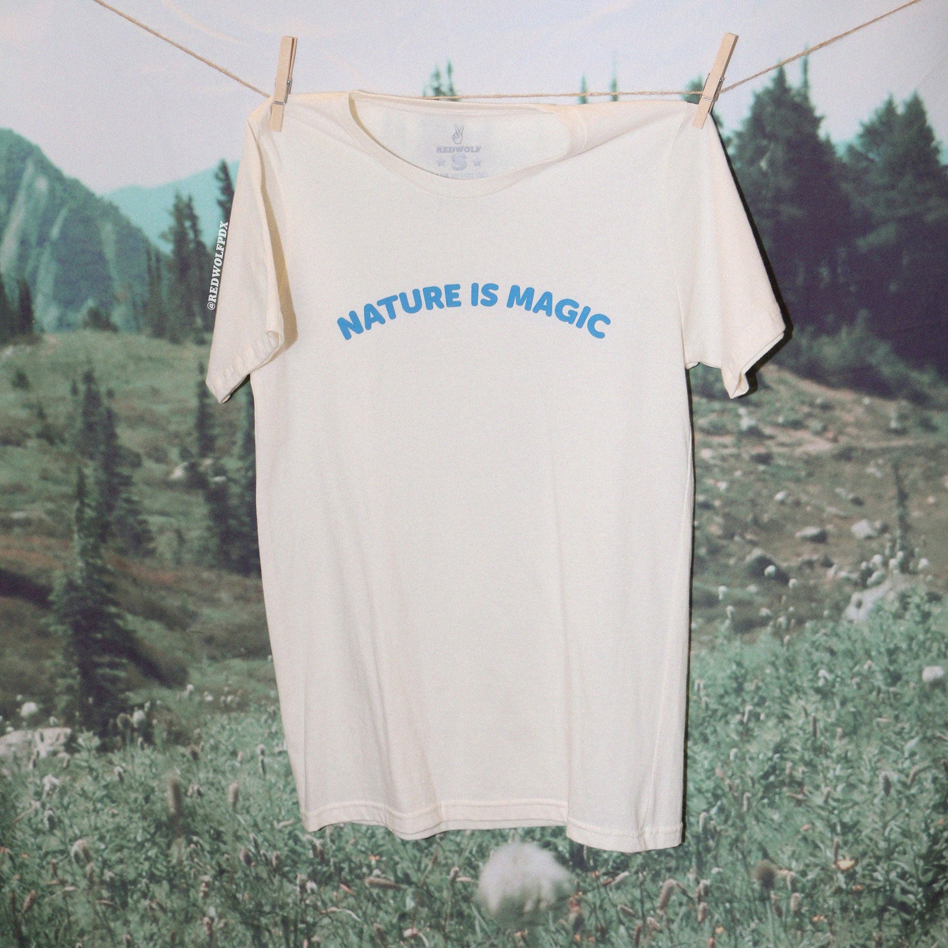 beige graphic tee with text 'NATURE IS MAGIC' in blue ink screen printed across chest hanging from clothing line against a nature backdrop