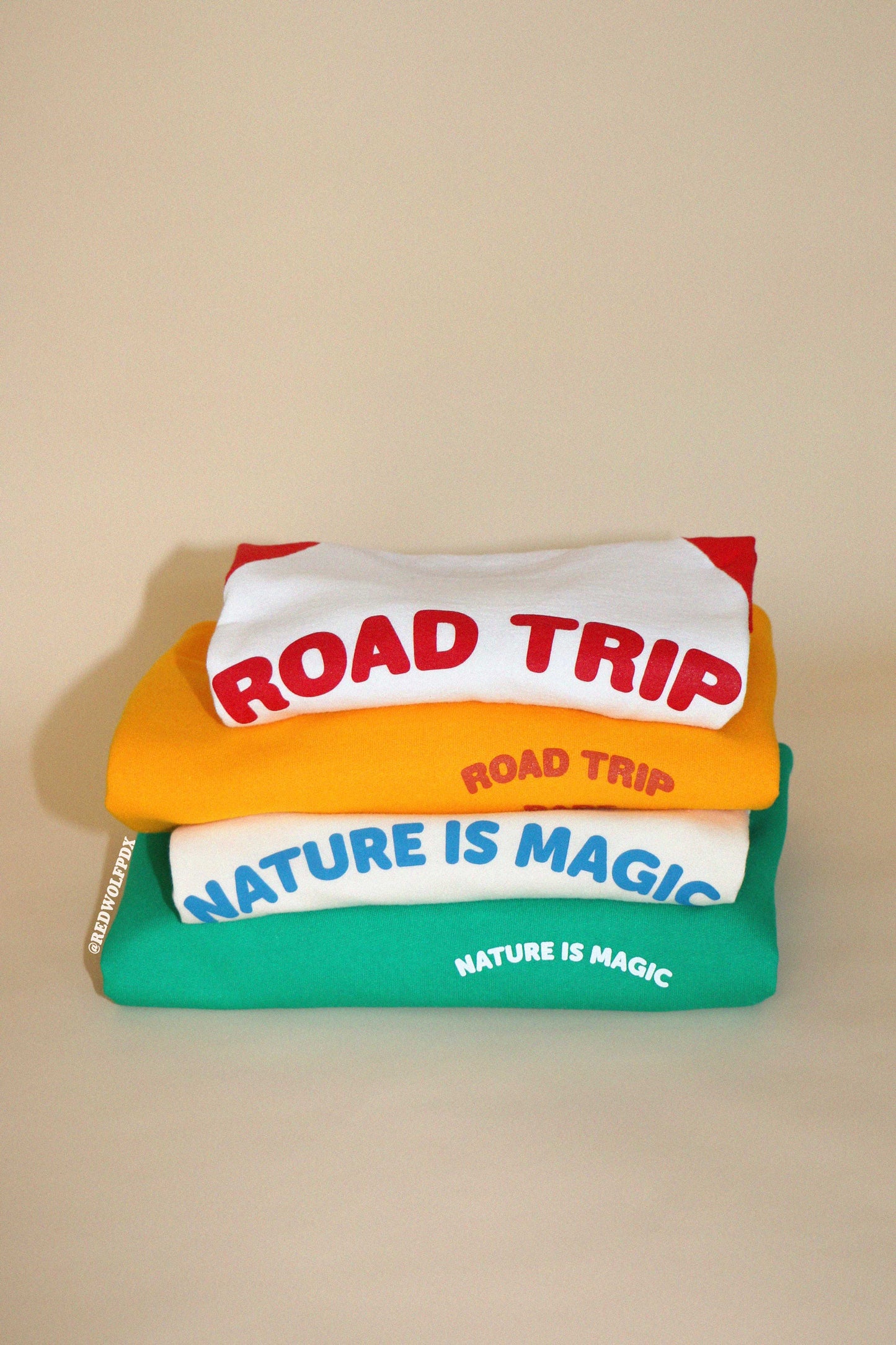Stack of colorful t-shirts with 'Road Trip' and 'Nature is Magic' text on a beige background