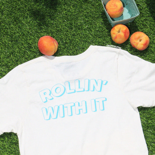 White graphic tee with the text 'ROLLIN' WITH IT' laying on grass with a carton of peaches