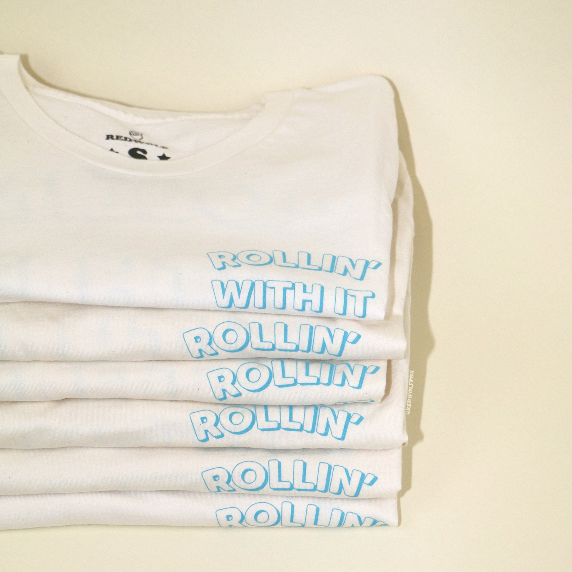 stack of beige white graphic tees with text 'ROLLIN' WITH IT' in blue ink on front corner of chest on a beige backdrop