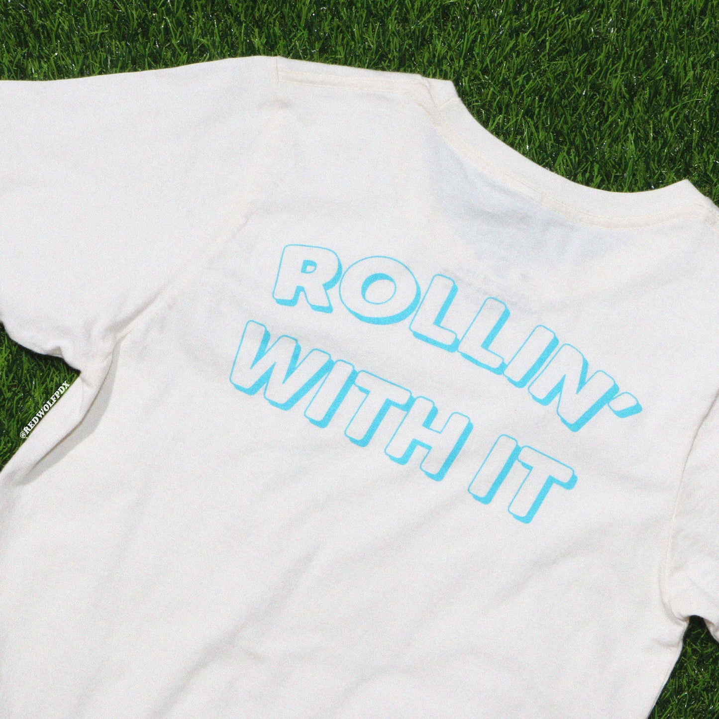 White graphic tee with the text 'ROLLIN' WITH IT' on green grass