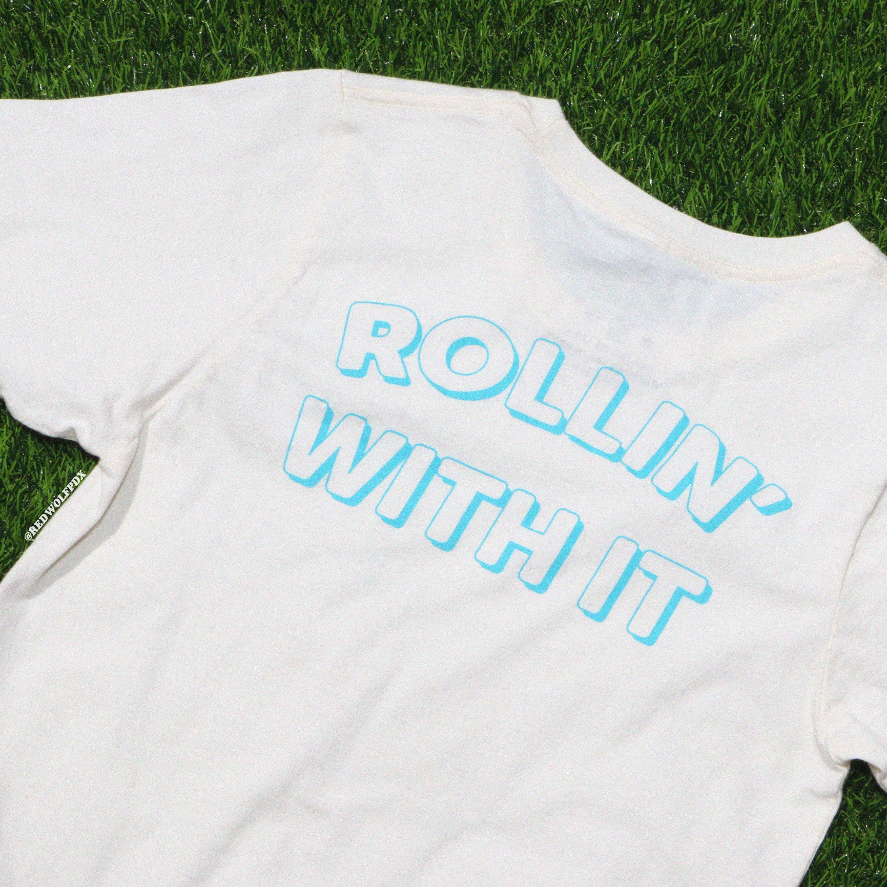 White graphic tee with the text 'ROLLIN' WITH IT' on green grass