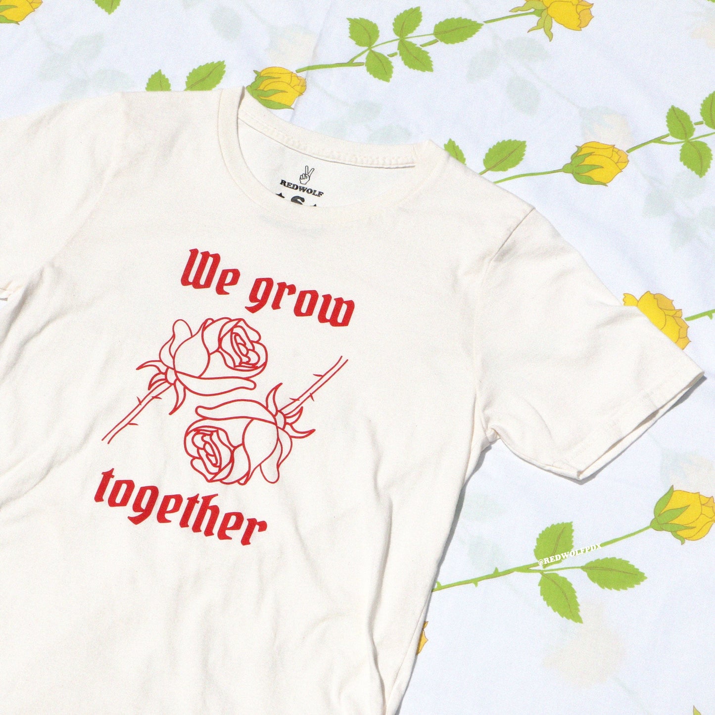 Beige graphic tee with text 'We grow together' and two red roses illustration in red ink screen printed on front hanging against a rose print backdrop