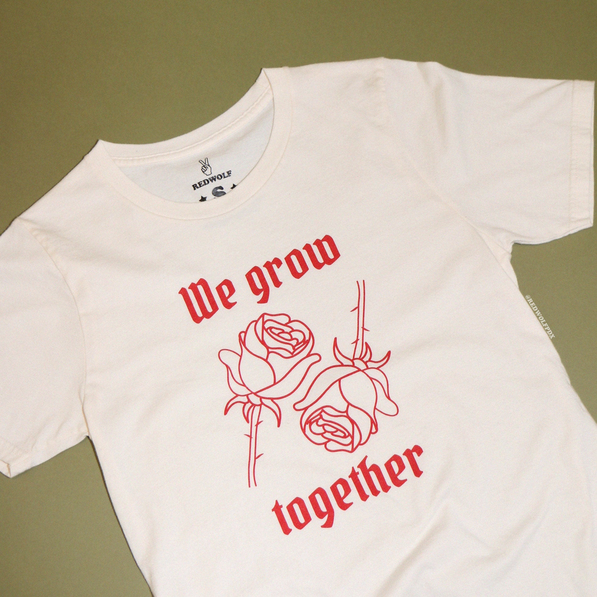 Beige graphic tee with text 'We grow together' and two red roses illustration in red ink screen printed on front against vintage green background