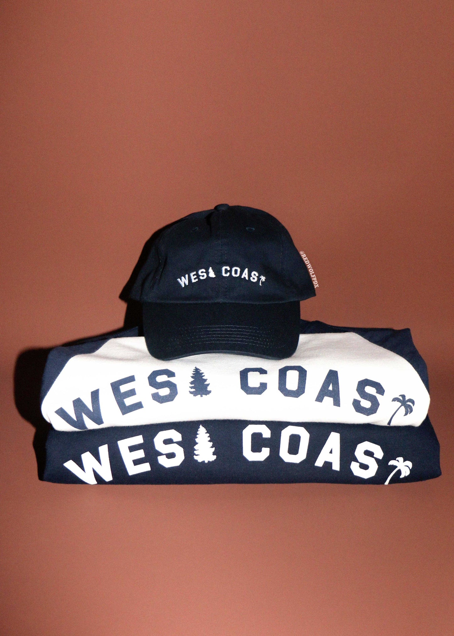 navy blue baseball cap stacked on white tee and navy blue crewneck sweatshirt all with 'WEST COAST' text on a brown background