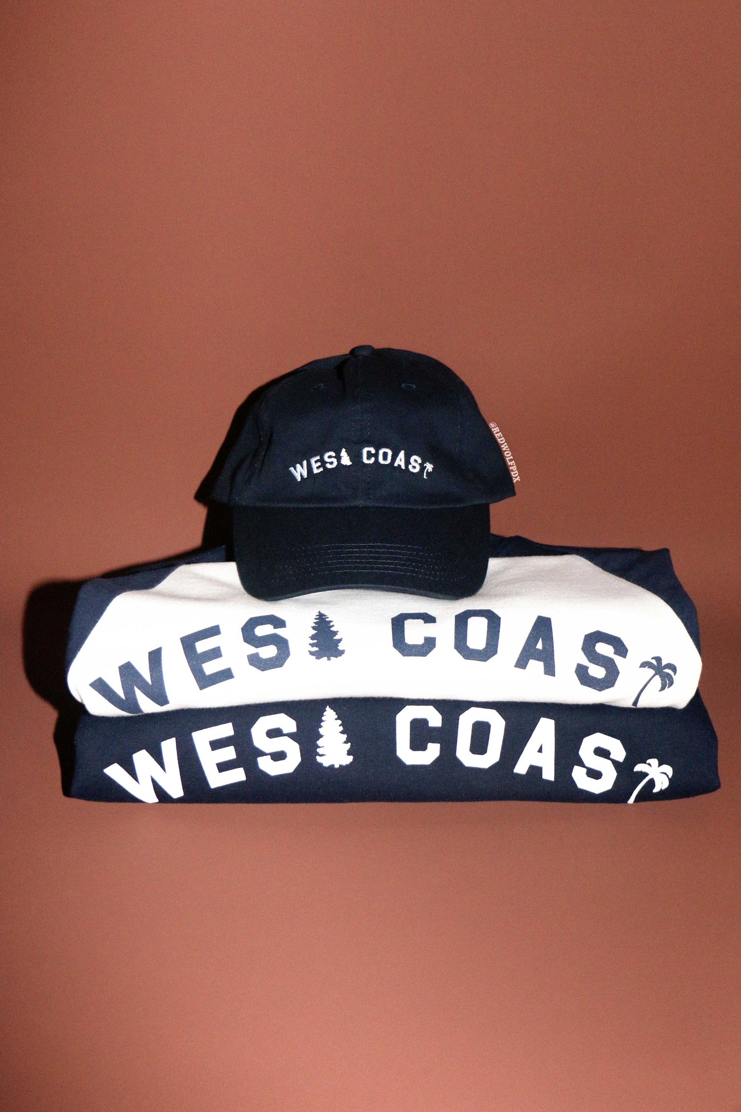 navy blue baseball cap stacked on white tee and navy blue crewneck sweatshirt all with 'WEST COAST' text on a brown background