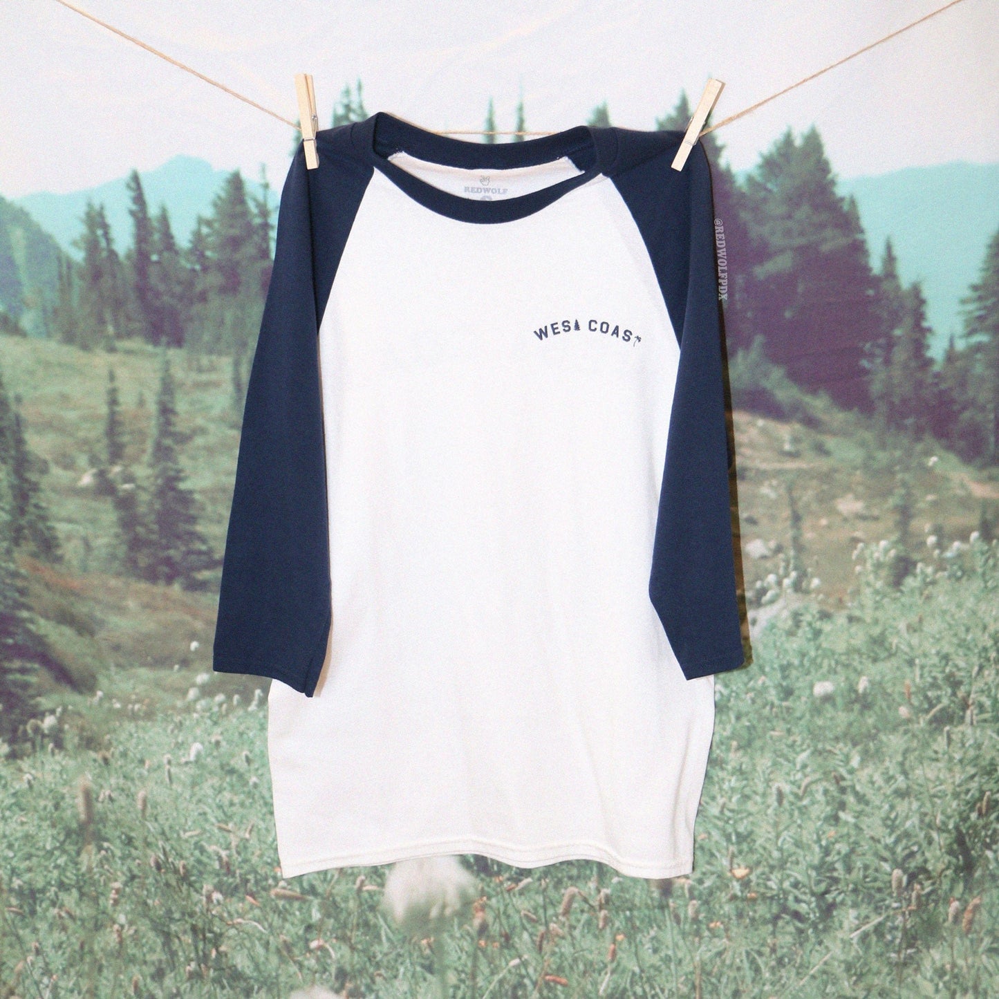 A navy blue and white raglan shirt with the text 'WEST COAST' printed across the chest with a nature backdrop