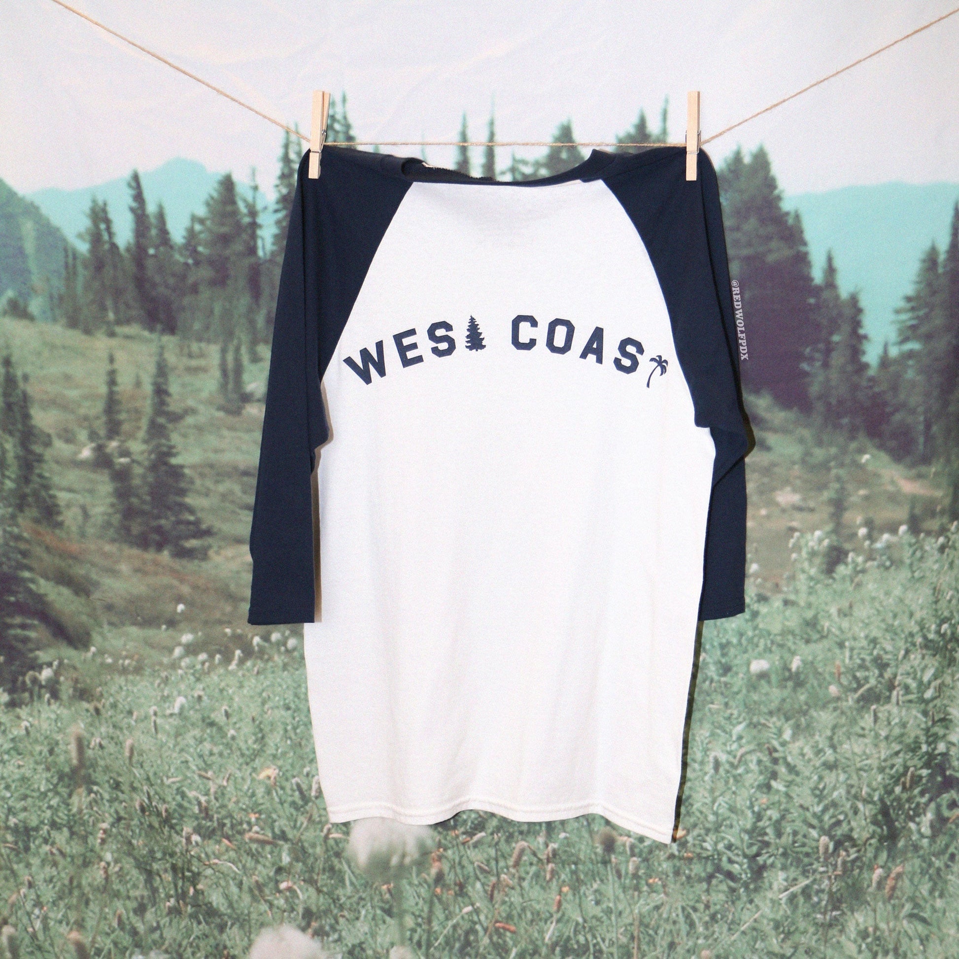 A navy and white raglan tee with the text 'WEST COAST' hanging on a wooden hanger against a natural and forest background.