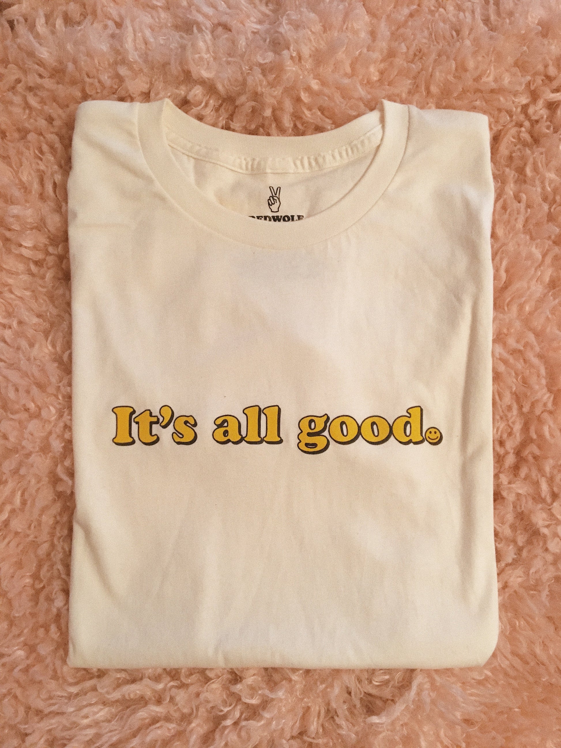beige folded graphic tee with text 'It's all good' with smiley face illustration in yellow ink screen printed across chest on a pink shaggy rug background