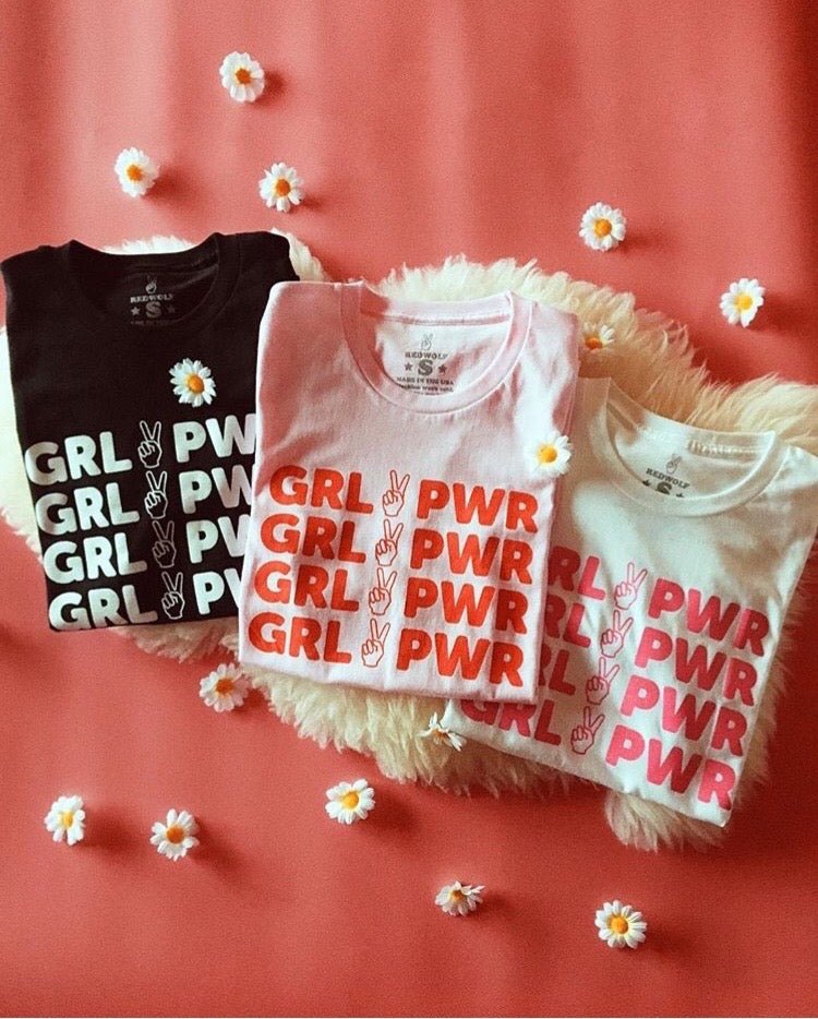 folded tees with text 'GRL PWR' with peace sign graphic screen printed across in multiple colors on faux fur rug on a pink background with daisies scattered across