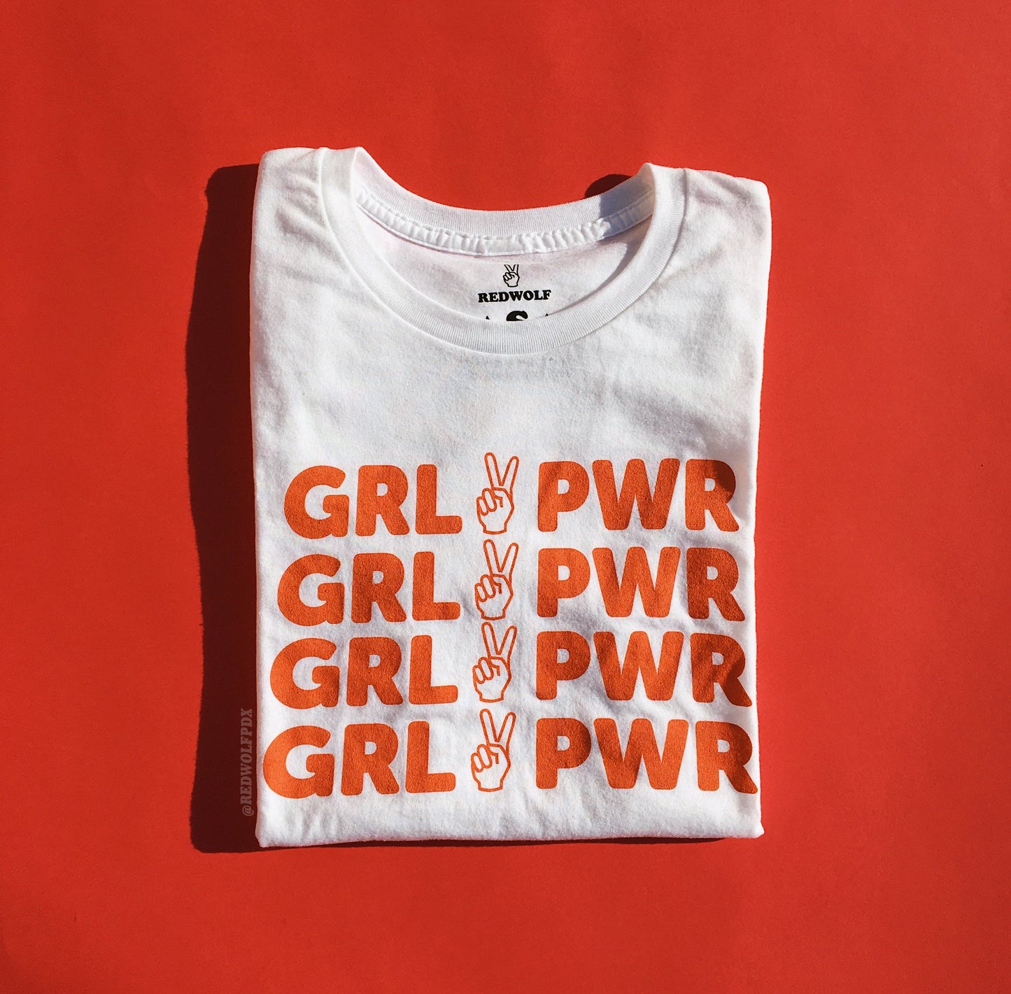 folded white graphic tee with orange red ink text 'GRL PWR' graphic across front against a vintage red backdrop