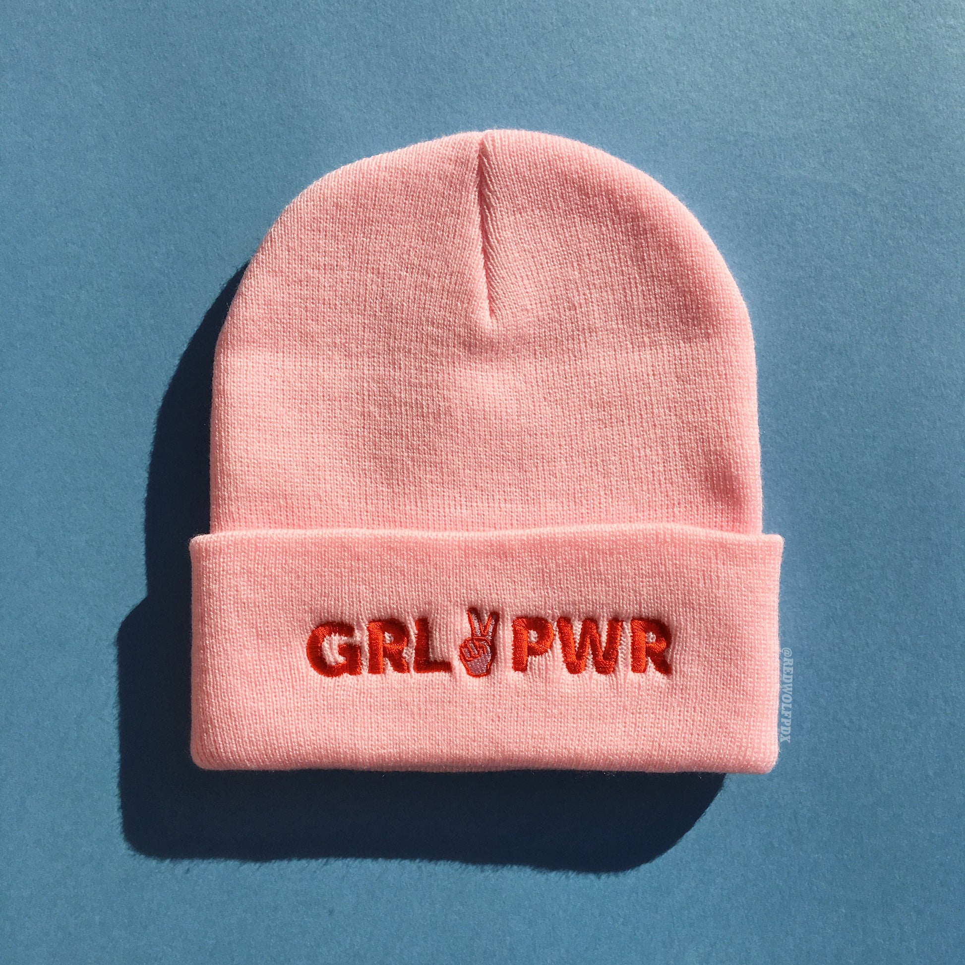 light pink beanie with text 'GRL PWR' in red thread embroidered across band of the beanie against a blue background