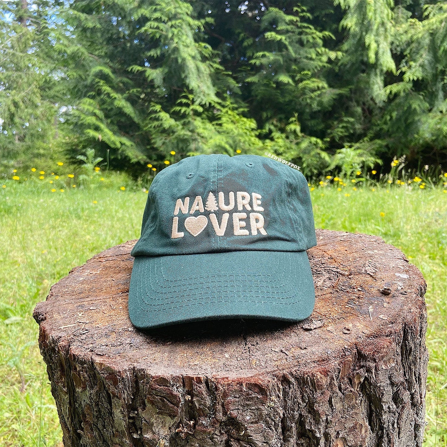 a forest green baseball cap with text 'NATURE LOVER' embroidered with beige thread across the front rested on a tree stump with a nature background