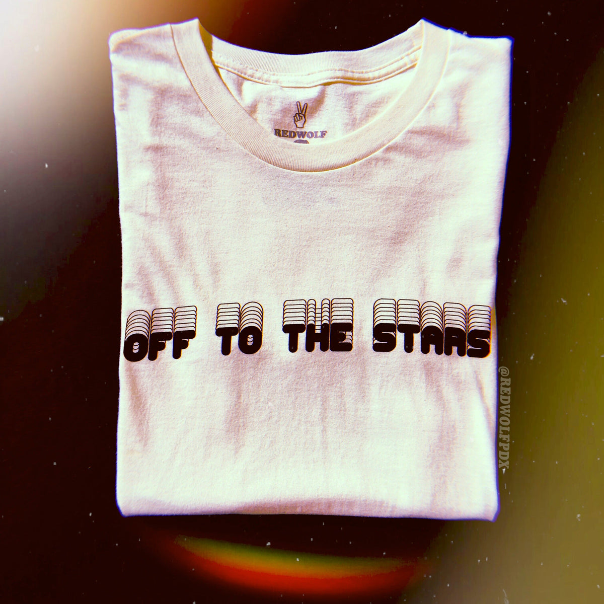 Folded white tee with text 'OFF TO THE STARS' on chest against space background