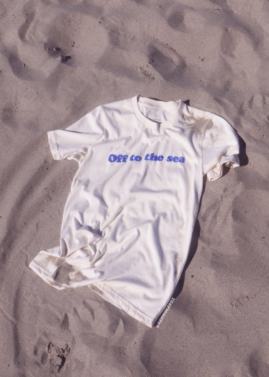 Beige white graphic tee with text 'Off to the sea' screen printed with raised blue ink across front sprawled out on sand