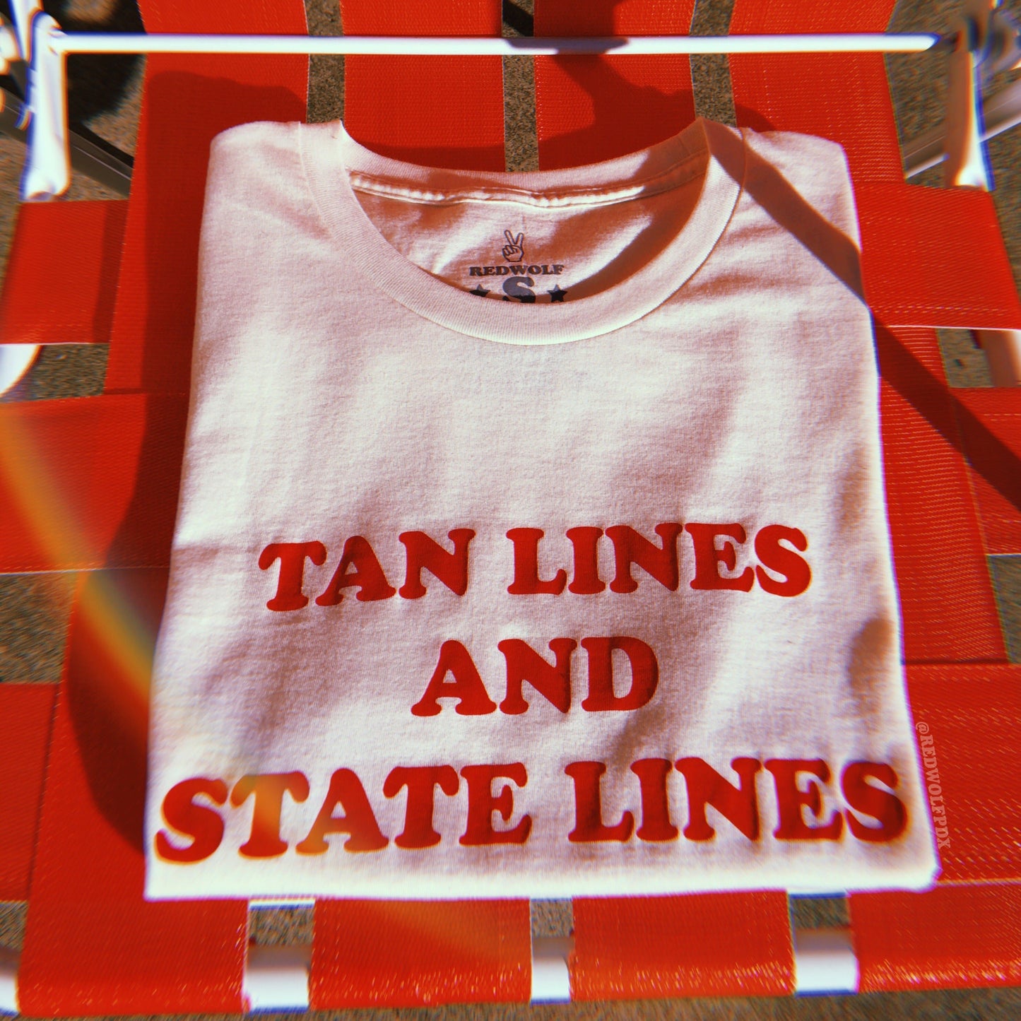 Folded beige graphic tee with text 'TAN LINES AND STATE LINES' in orangey red screen printed on chest rested on red lawn chair.