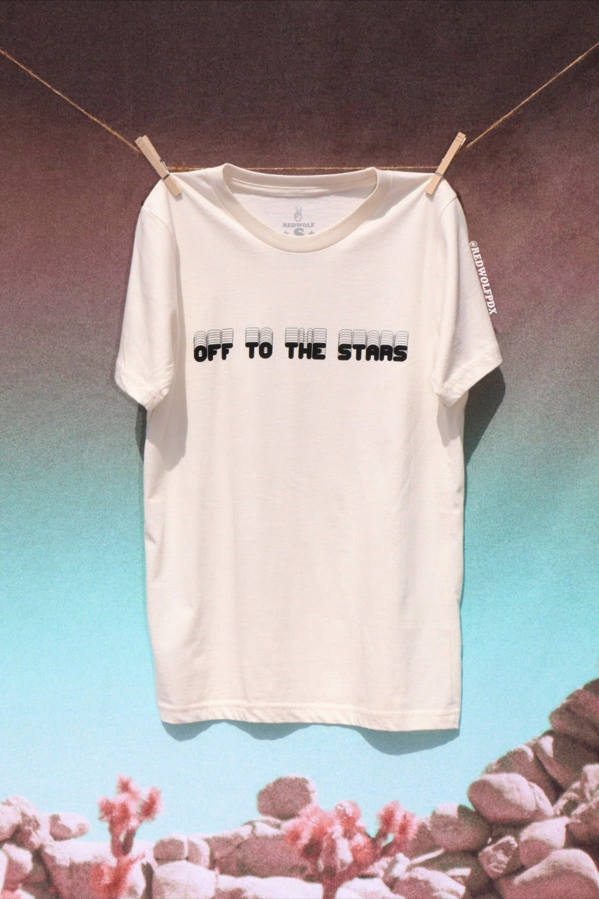 White t-shirt with text 'Off to the Stars' hanging on a clothesline with a space-themed background.