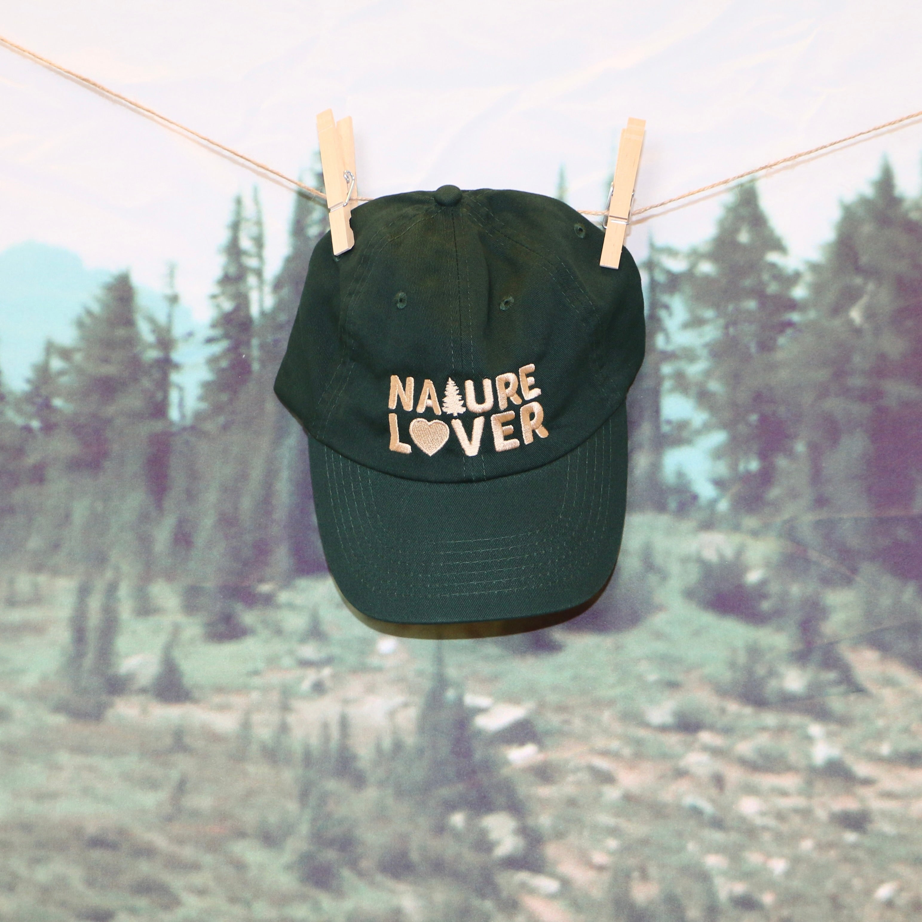 forest green baseball cap with text 'NATURE LOVER' embroidered in beige thread across the front being held by wood clothes pins laundry line against a nature backdrop