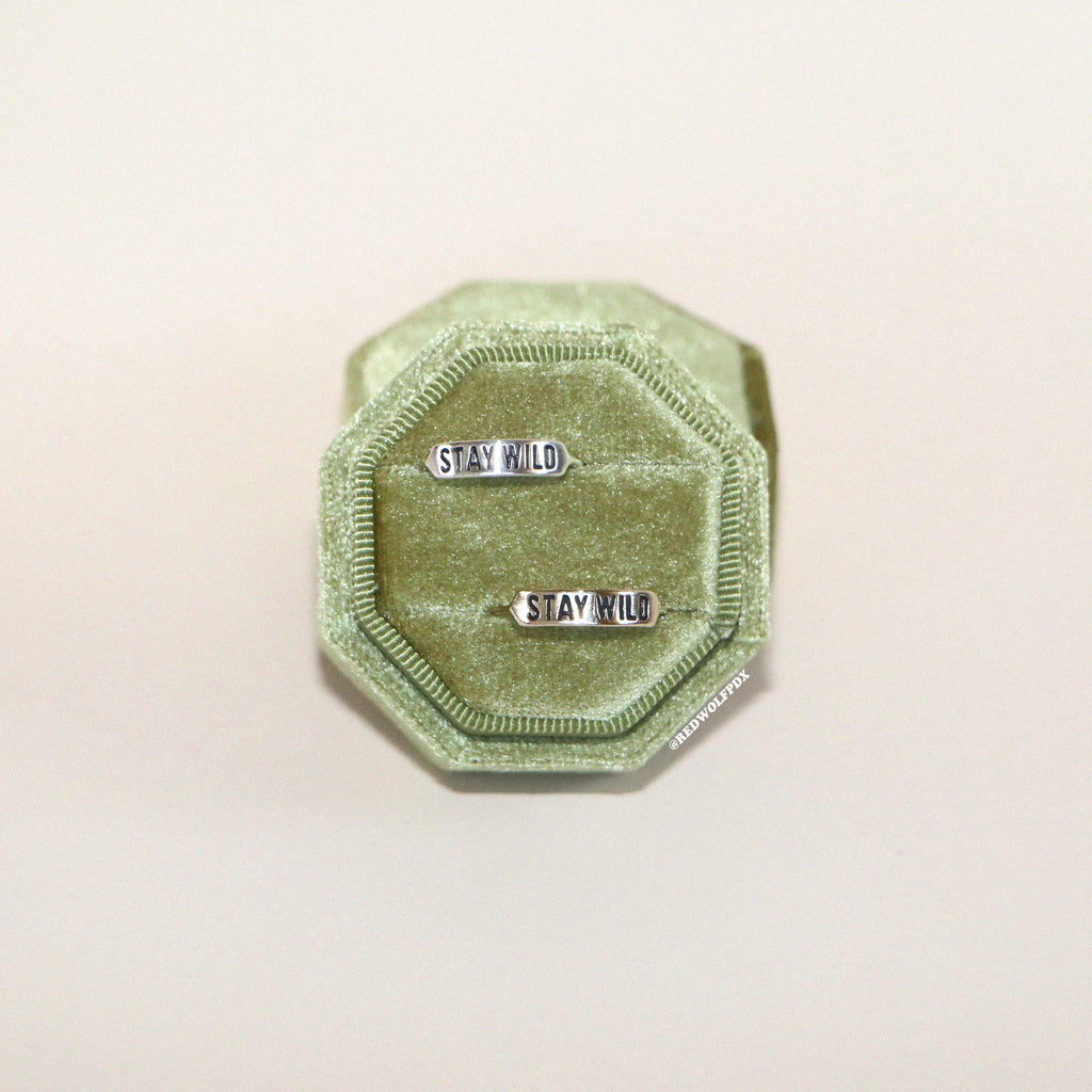 A sterling silver  ring and brass ring with  text 'STAY WILD' engraved on the bands, displayed a green velvet ring box on a white background.