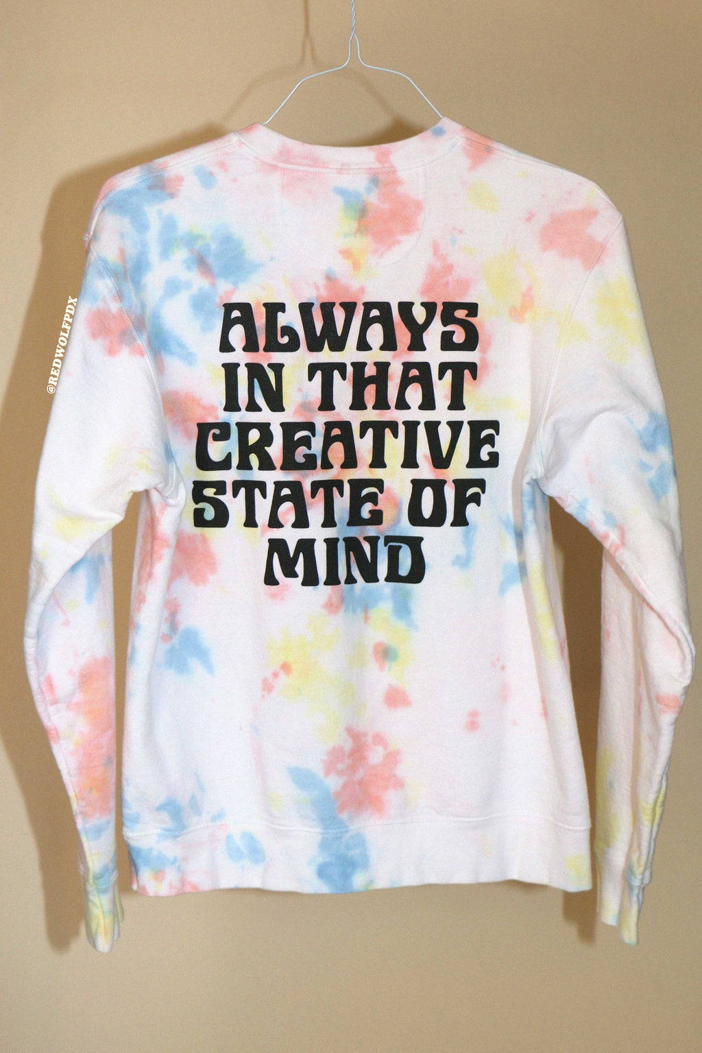 Colorful tie-dye crewneck sweatshirt with text 'ALWAYS IN THAT CREATIVE STATE OF MIND' across the back against a beige background