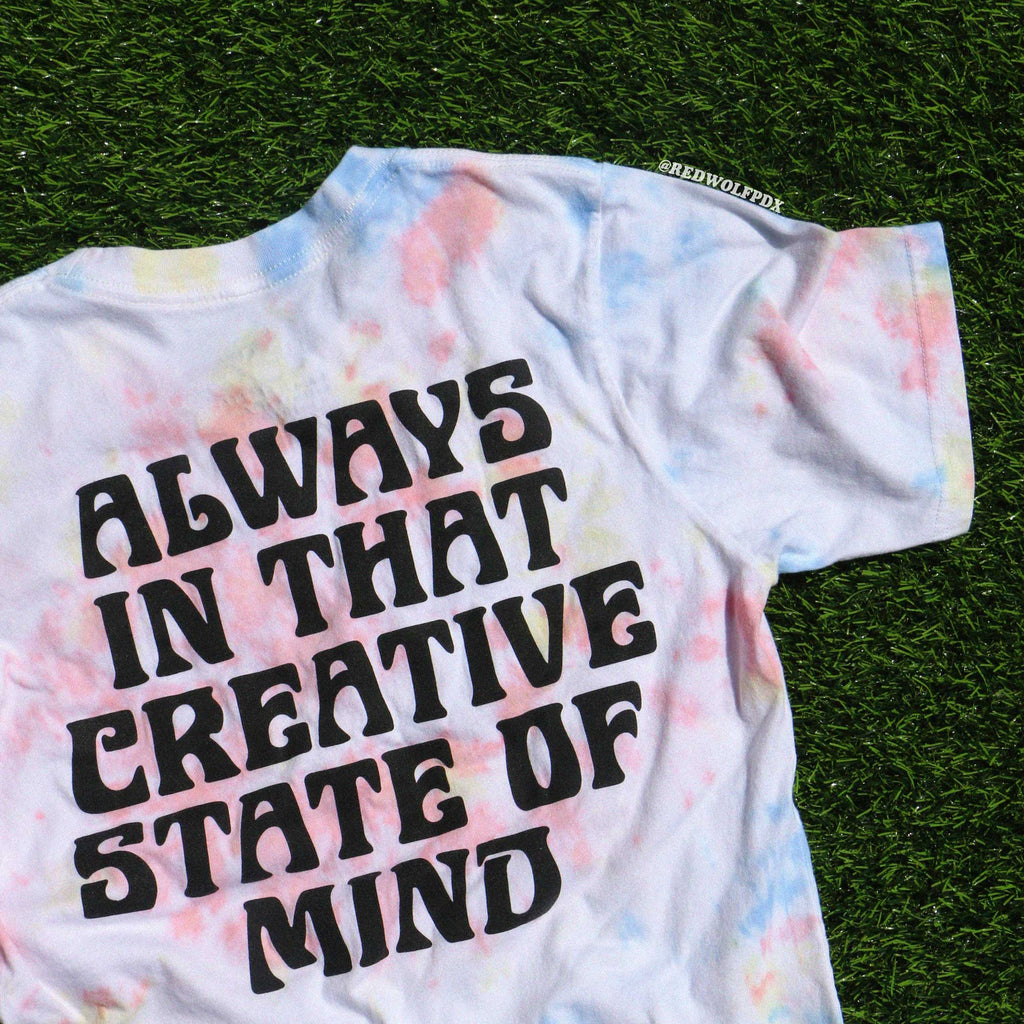T-shirt with 'Always in that creative state of mind' text on a tie dye background, laid flat on a green surface.