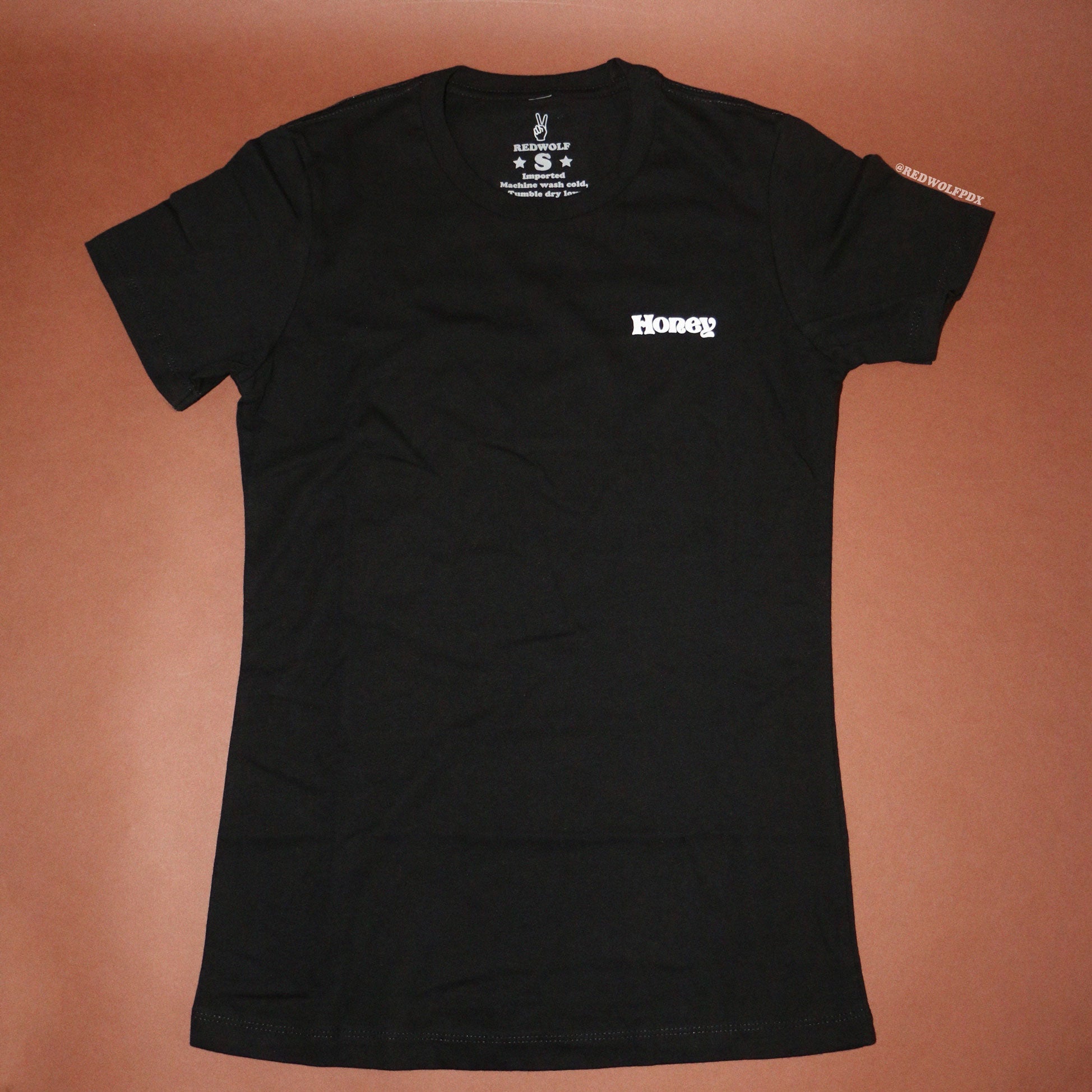 Black fitted tee with text 'Honey' on corner of chest against brown backdrop