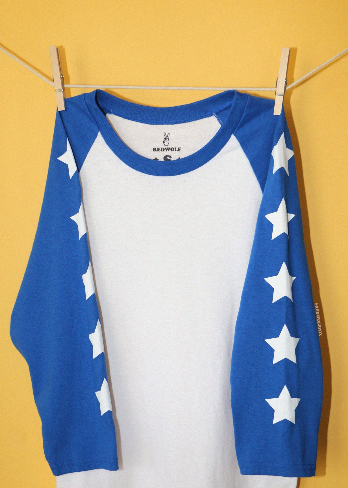 A raglan-style long-sleeve shirt with a white body and royal blue sleeves, decorated with white stars down the sleeves, hanging from a clothesline against a solid yellow background.