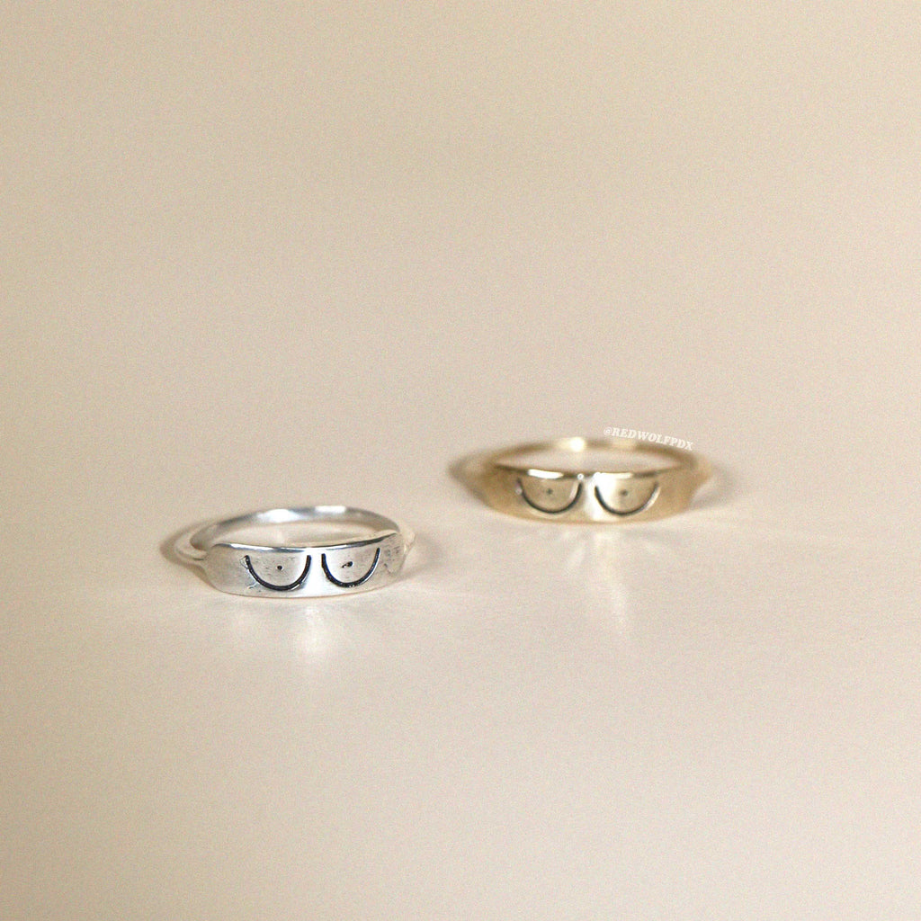 Two rings, one made of sterling silver and the other of brass, each with an engraved illustration of boobs across it , displayed against a neutral background.