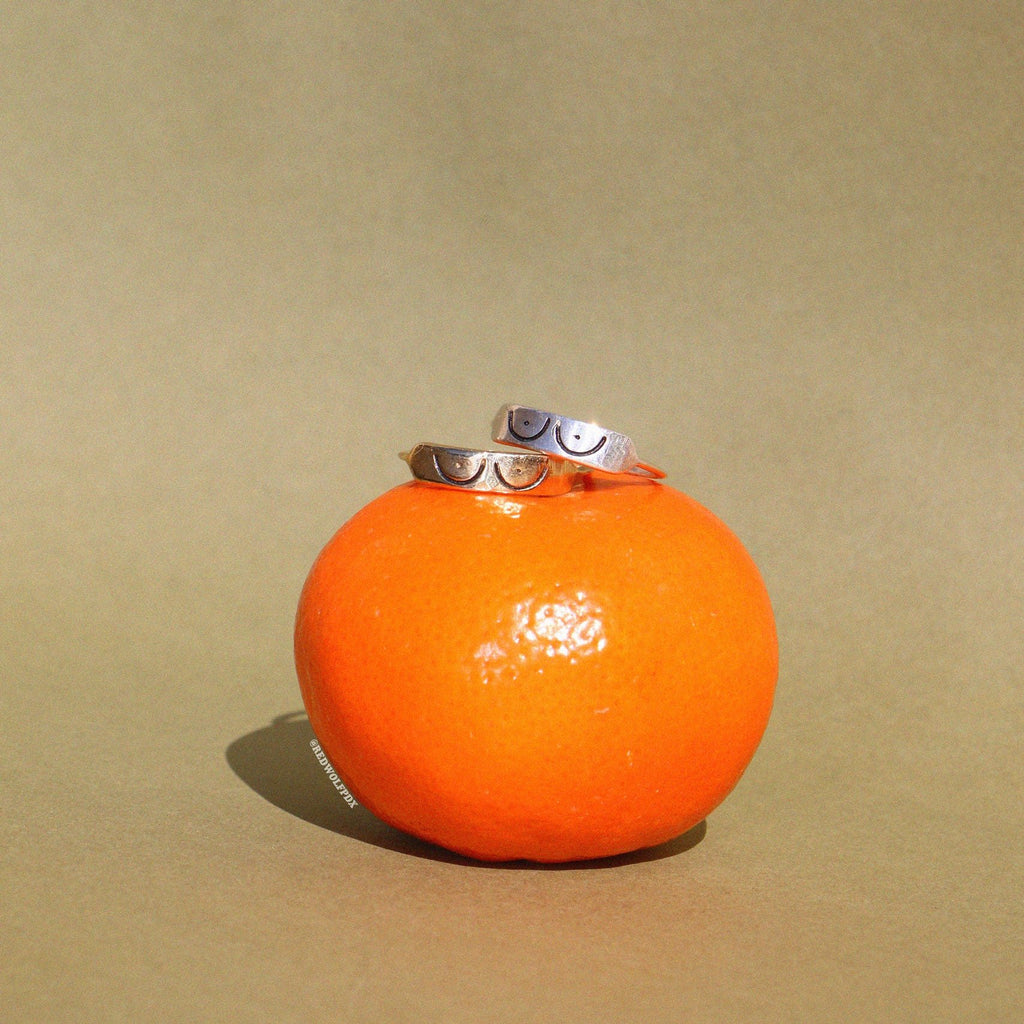 This image features a striking piece of jewelry known as the "Boobs Ring." Displayed on a vibrant orange fruit, which adds a pop of color against the neutral backdrop, the ring is  designed with a 3D etching of a boobs illustration.