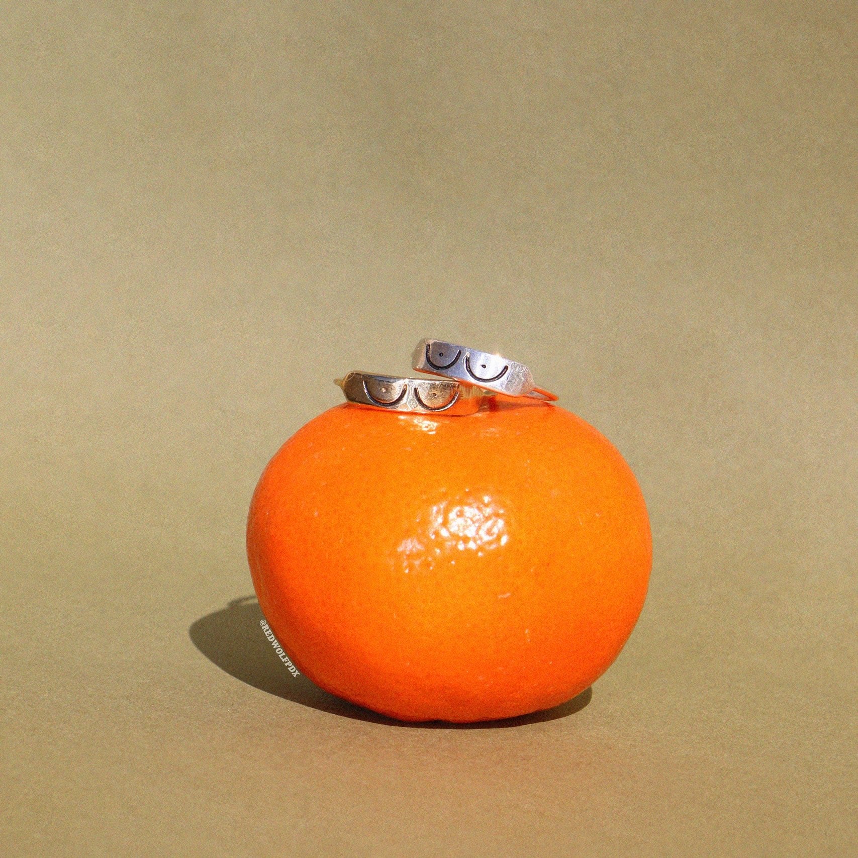 This image features a striking piece of jewelry known as the "Boobs Ring." Displayed on a vibrant orange fruit, which adds a pop of color against the neutral backdrop, the ring is  designed with a 3D etching of a boobs illustration.