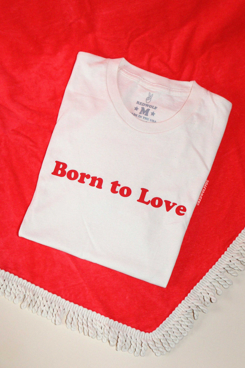 A white crew neck t-shirt with the red graphic 'Born to Love' printed across the front, displayed on a red heart background.