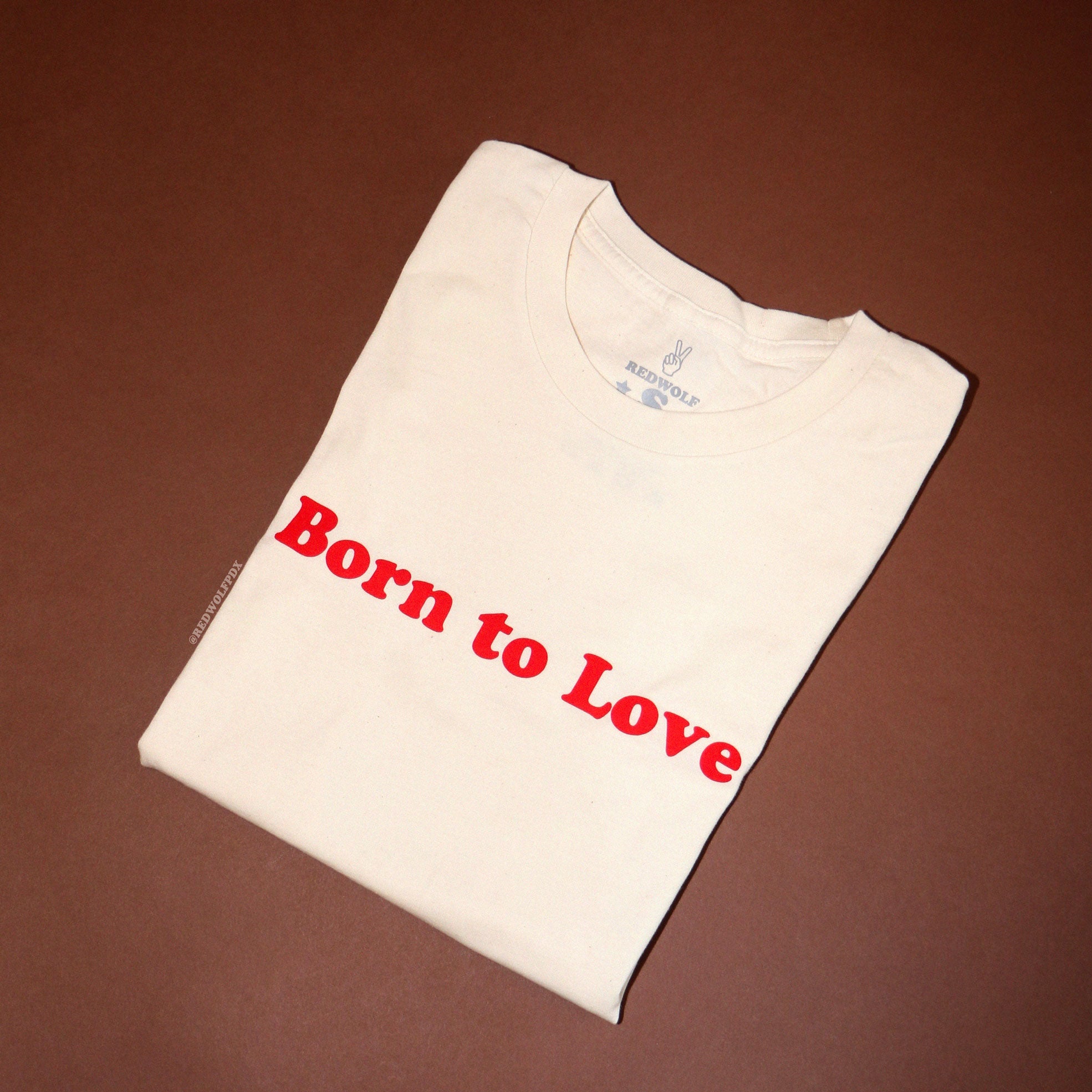 Folded white graphic tee with the text 'Born to Love' across the chest against brown paper backdrop