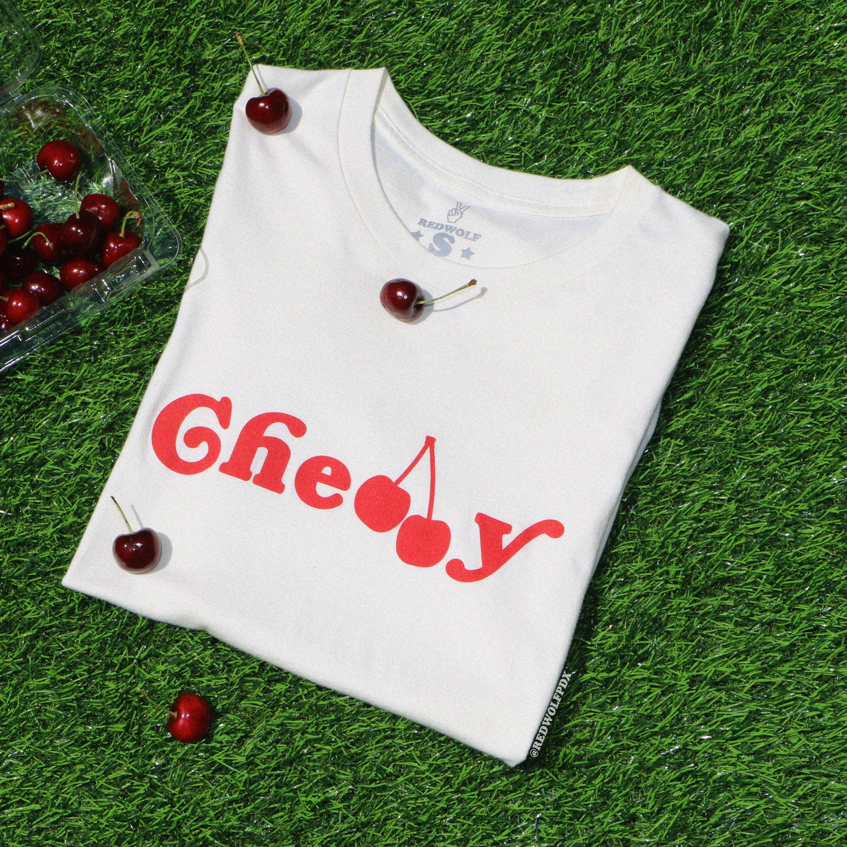 Folded beige white graphic tee with text 'Cherry' screen printed across in red ink on green grass with a carton of scattered cherries