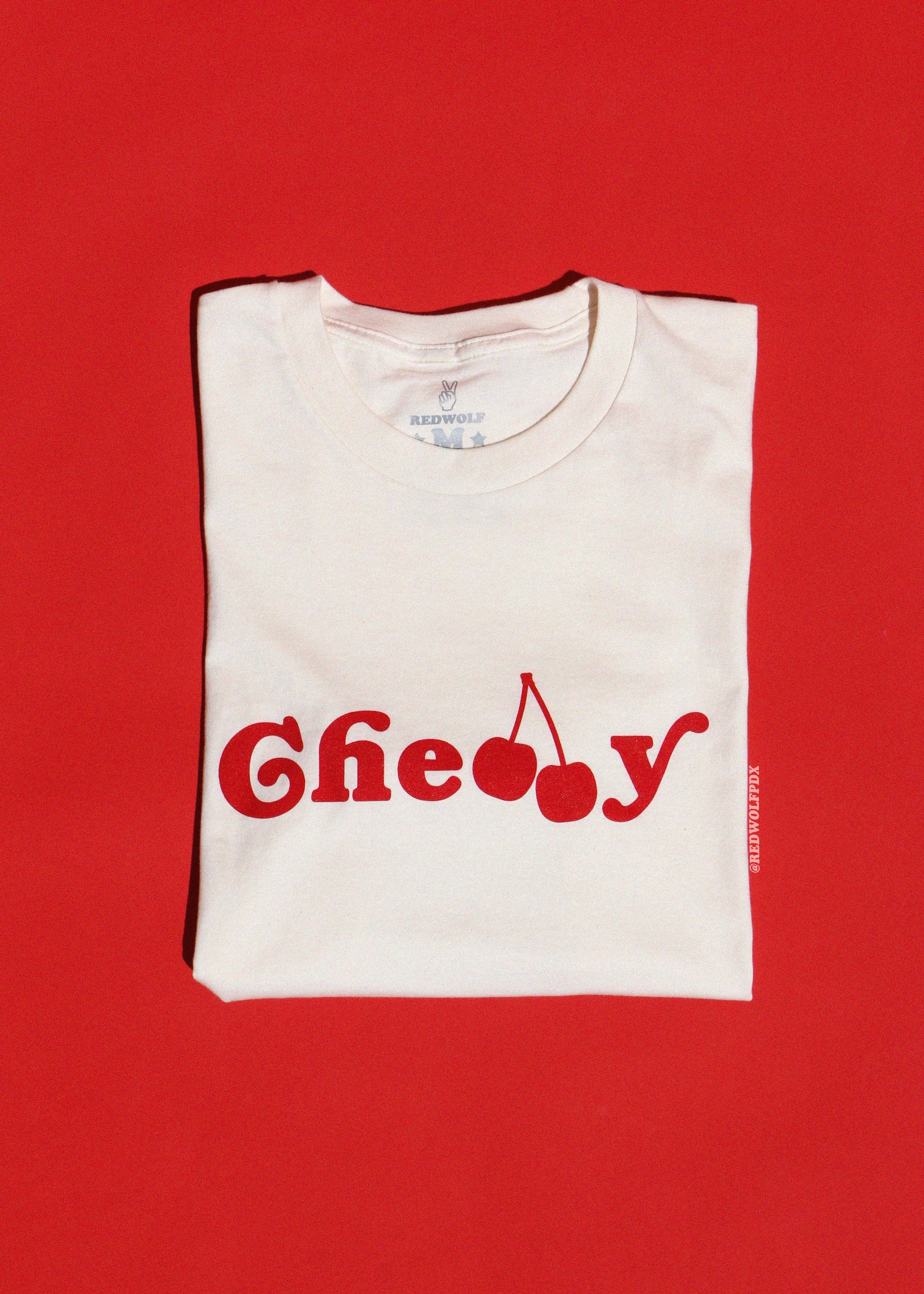 Folded white t-shirt with red text and cherry graphic on a red background