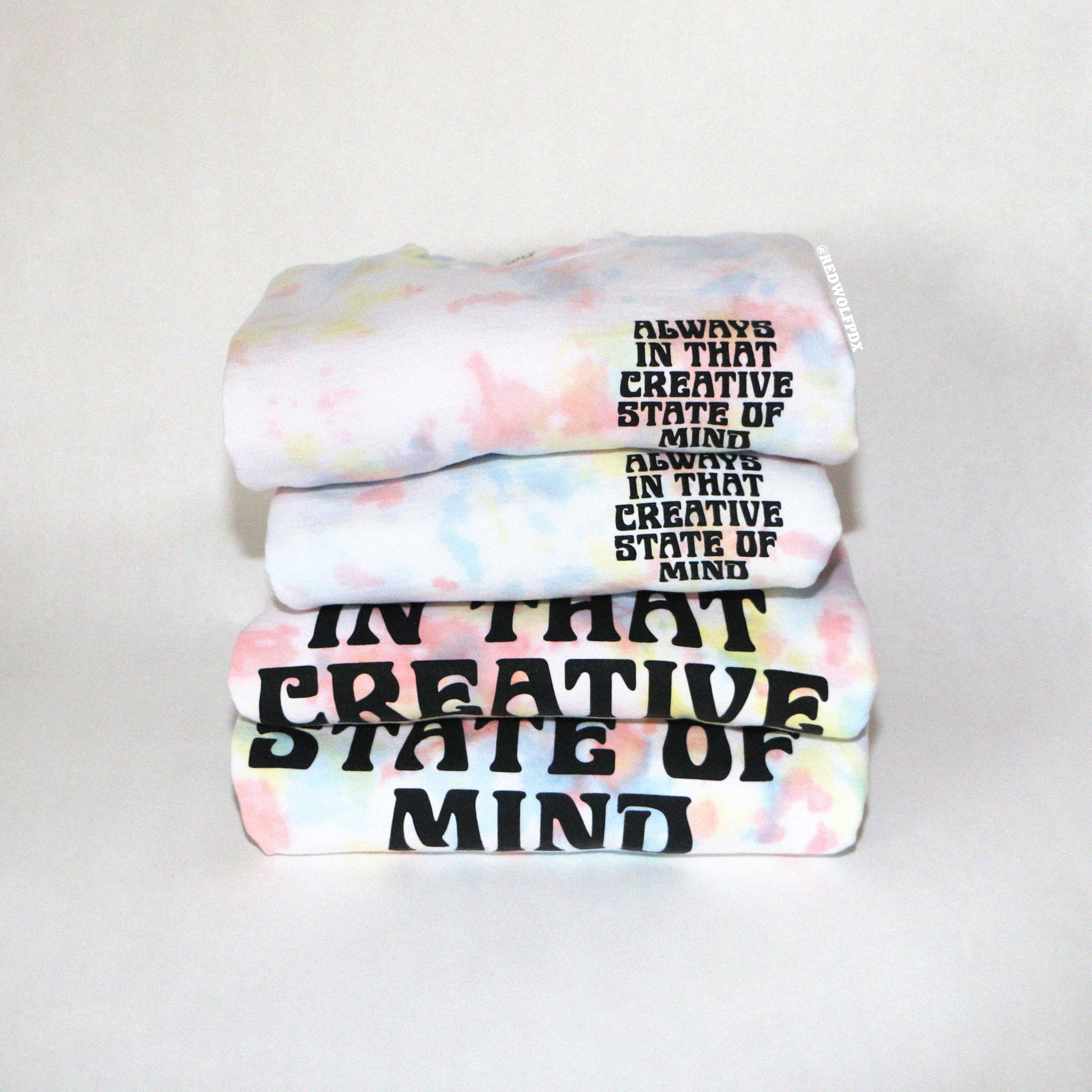Stack of tie dye crewnecks  with text 'ALWAYS ON THAT CREATIVE STATE OF MIND' screen printed on front and back on a white background