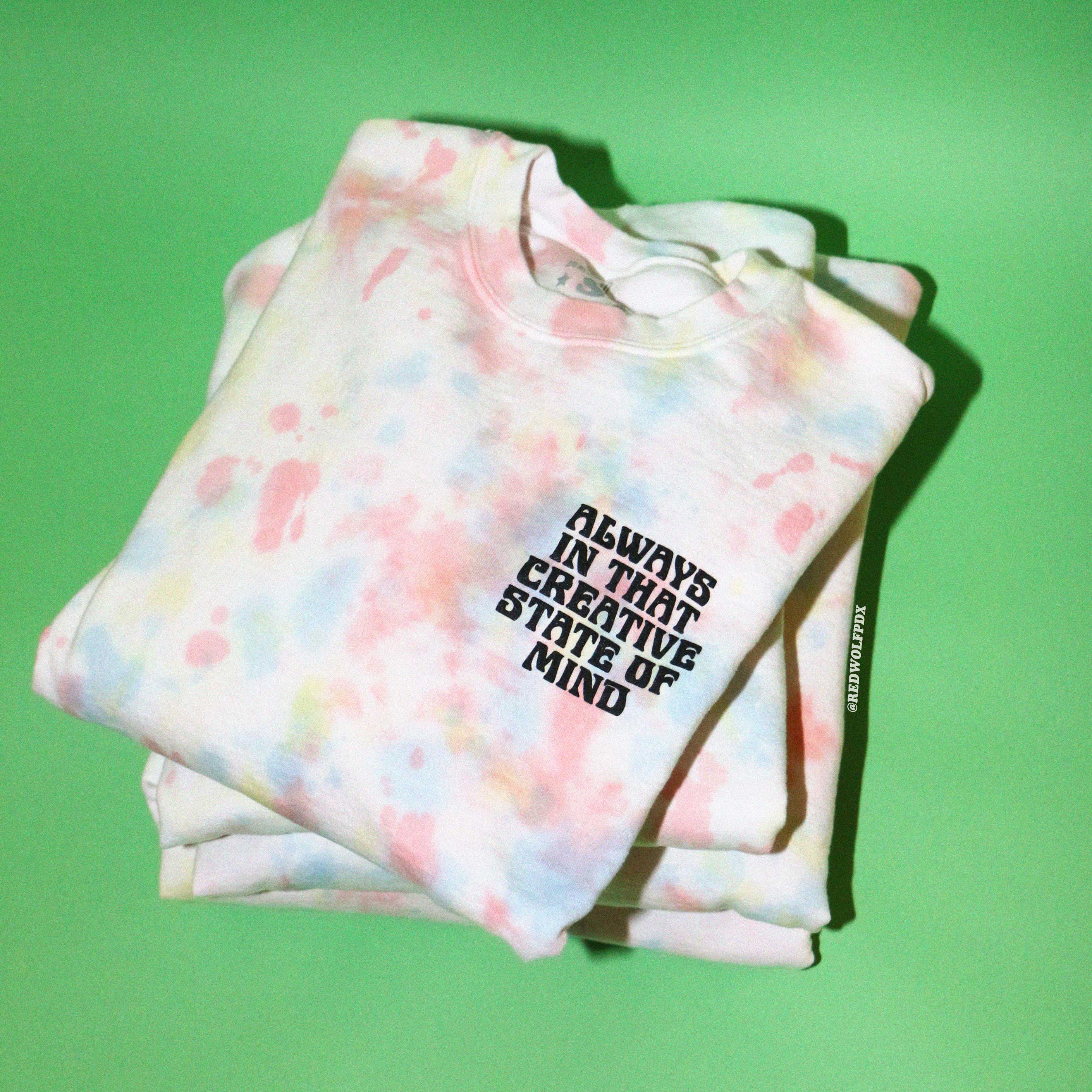 Stack of pastel tie-dye crewneck sweatshirts  with text 'ALWAYS IN THAT CREATIVE STATE OF MIND' on a green background