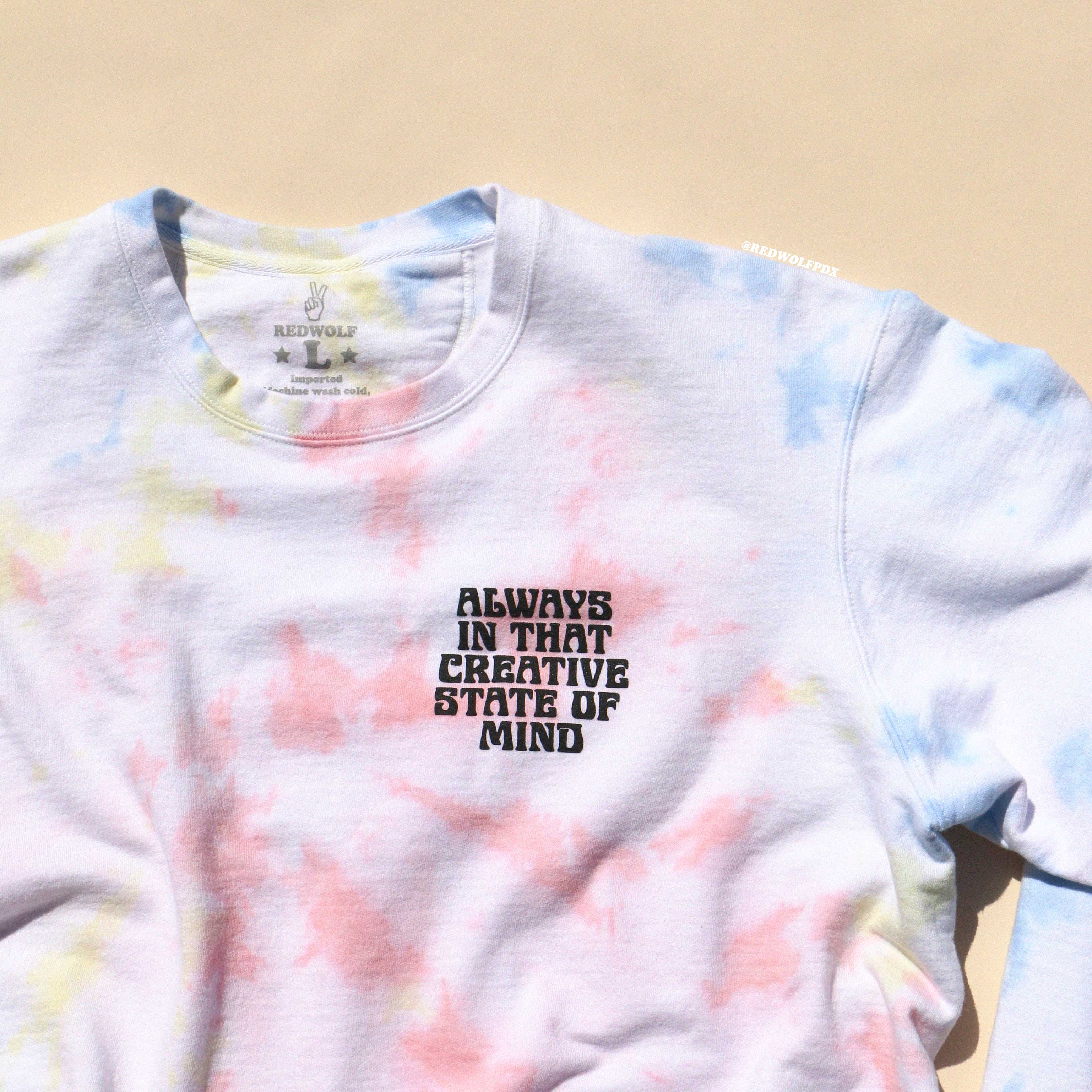 A tie-dye crewneck sweatshirt with a colorful spiral pattern and text graphic that reads 'ALWAYS IN THAT CREATIVE STATE OF MIND' on the front.
