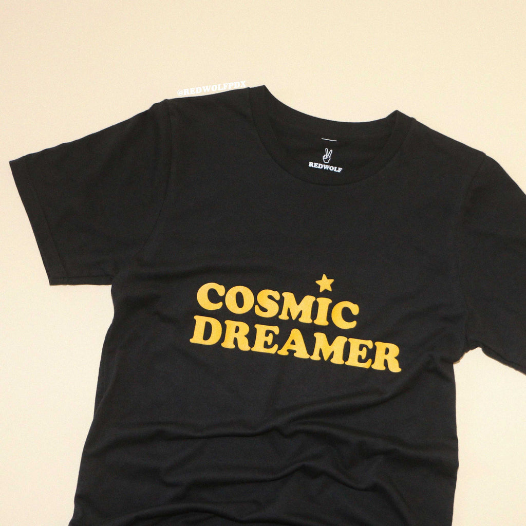 black graphic tee with text 'COSMIC DREAMER' in yellow ink screen printed across chest against a beige backdrop