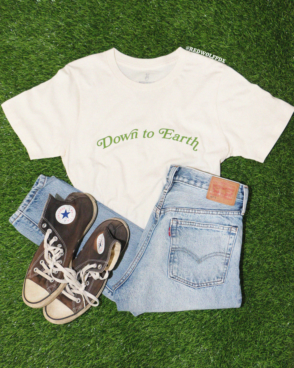 Beige graphic tee with text 'Down to Earth'  in green ink laid out in a flat lay with blue jeans and sneakers on green grass