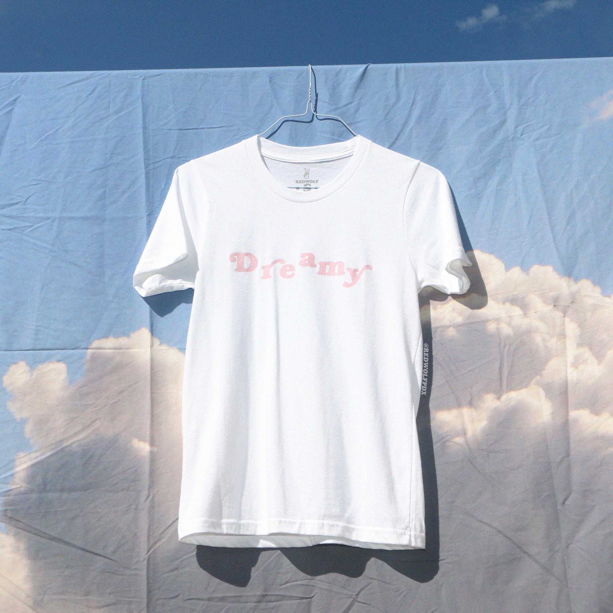 White graphic tee with text 'Dreamy' in bubble gum pink screen printed across the chest hanging on a sky backdrop