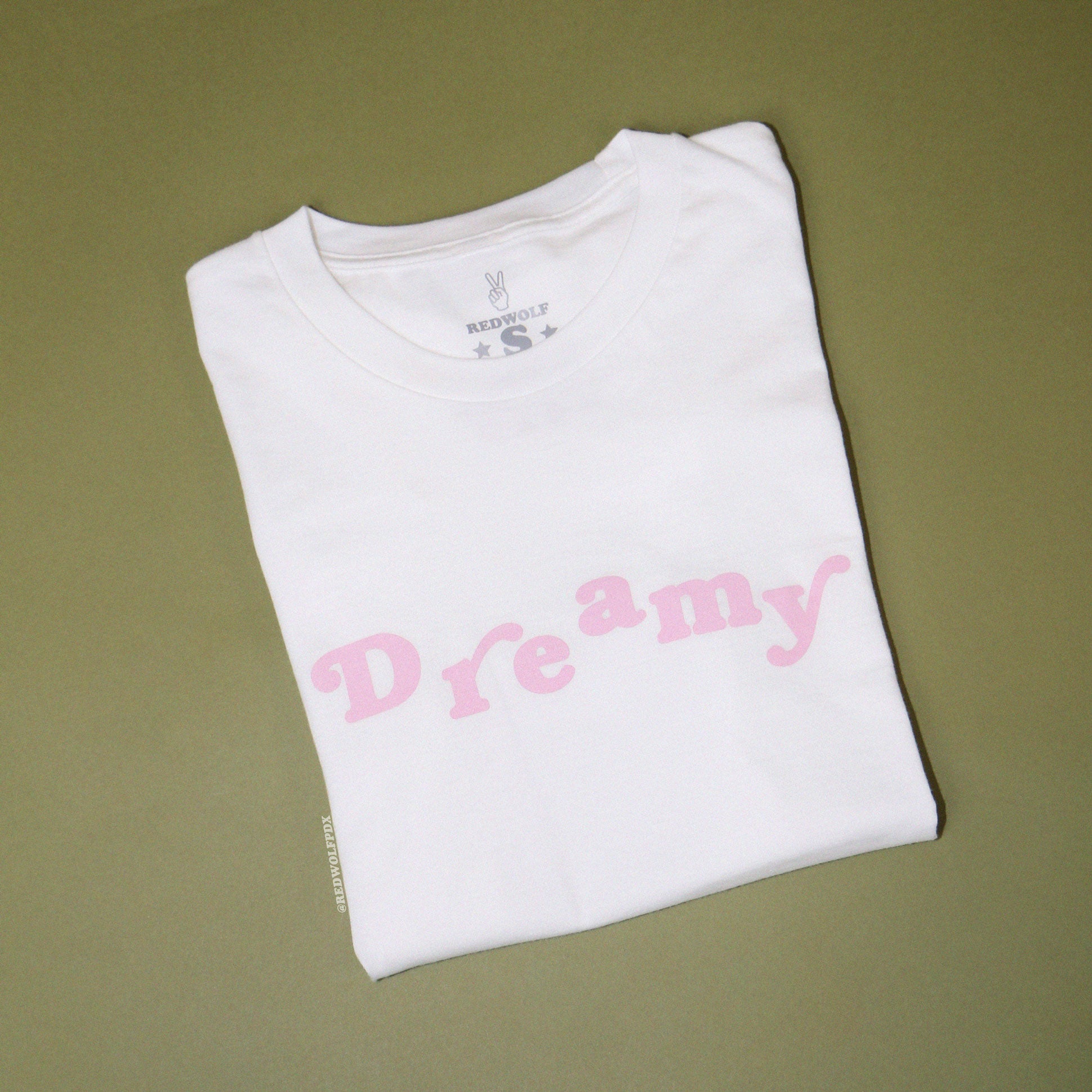 Folded white graphic tee with text 'Dreamy' in baby pink ink screen printed on front against a vintage green backdrop