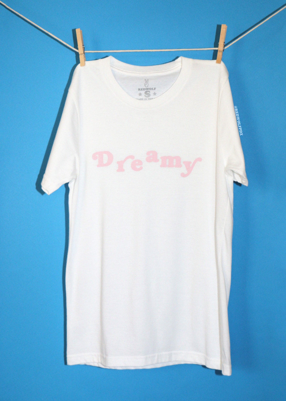 A white short-sleeve t-shirt with the word 'Dreamy' printed in pink across the chest, hanging from a clothesline with wooden clothespins against a solid blue background.